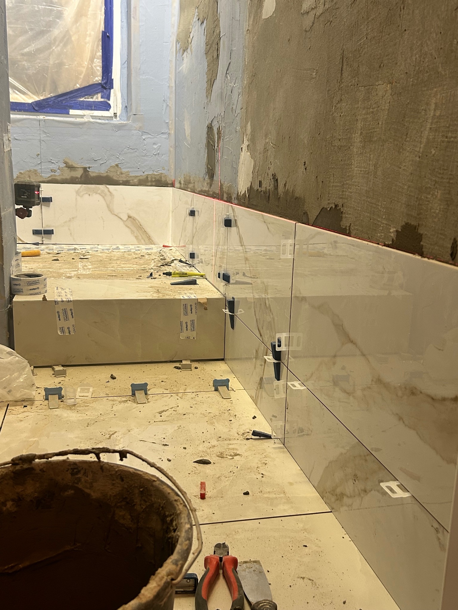 Układanie płytek imitujących marmur na ścianie w łazience, widoczne narzędzia glazurnicze, laserowy poziom, niezabezpieczone ściany i okno.