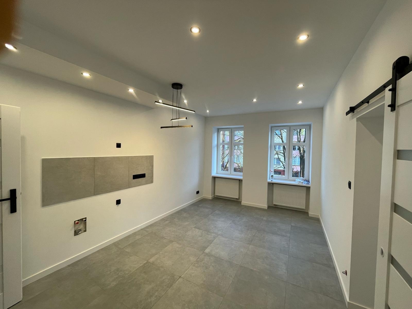 Wykończone wnętrze pokoju z szarą podłogą, białymi ścianami, nowoczesnym oświetleniem i elementami dekoracyjnymi na ścianie. Widoczne okna i drzwi przesuwne.