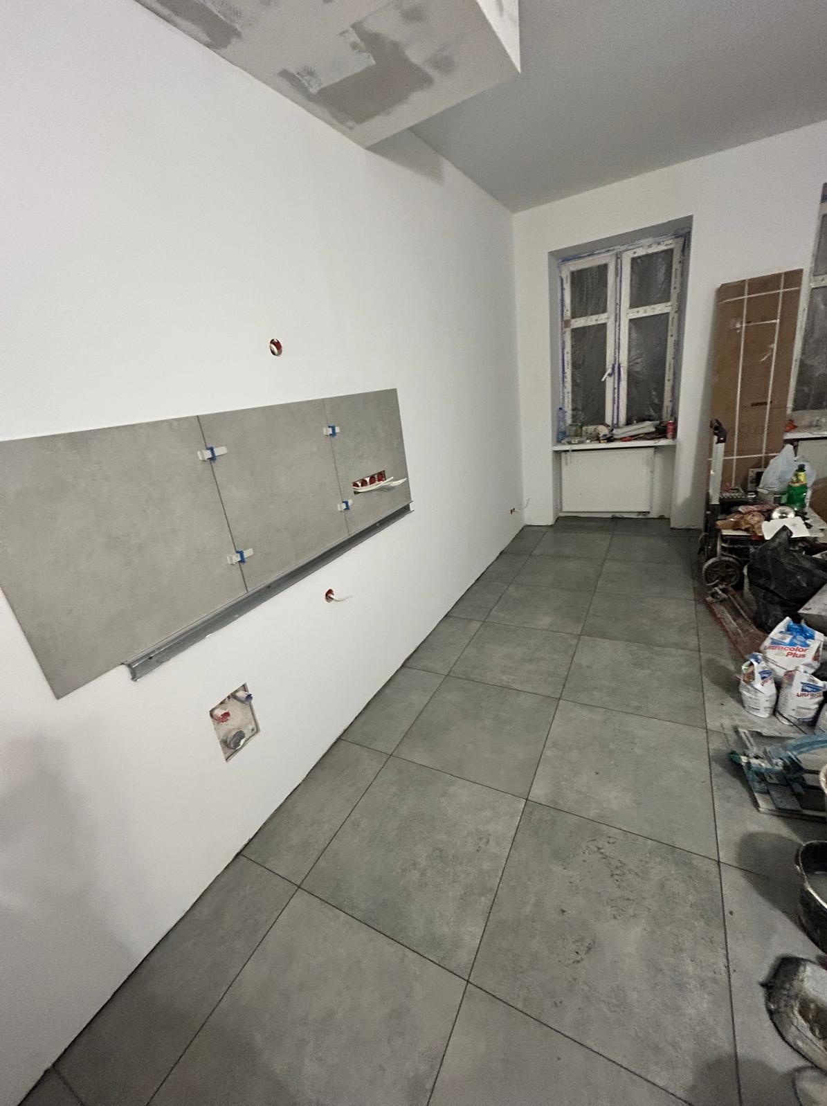 Szare płytki podłogowe i fragment ściany z zamontowanymi płytkami, widoczne krzyżyki dystansowe. W tle okno i materiały budowlane.