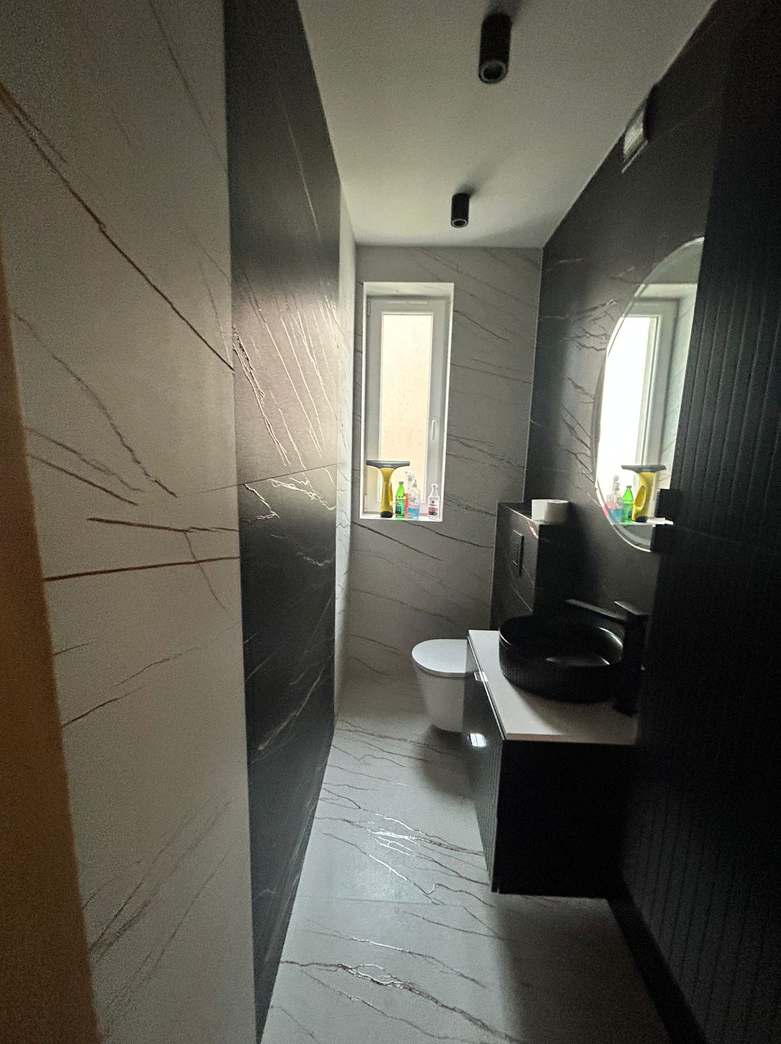 Nowoczesna łazienka z czarno-białymi płytkami imitującymi marmur, okrągłym lustrem i czarną umywalką nablatową. Minimalistyczny design z akcentami kolorystycznymi na parapecie.