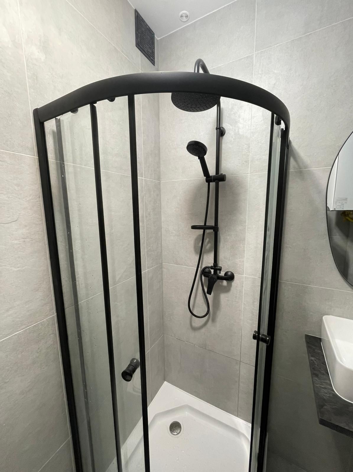 Nowoczesna kabina prysznicowa z czarną armaturą i szklanymi ściankami na tle szarych płytek w wyremontowanej łazience. Minimalistyczny design.