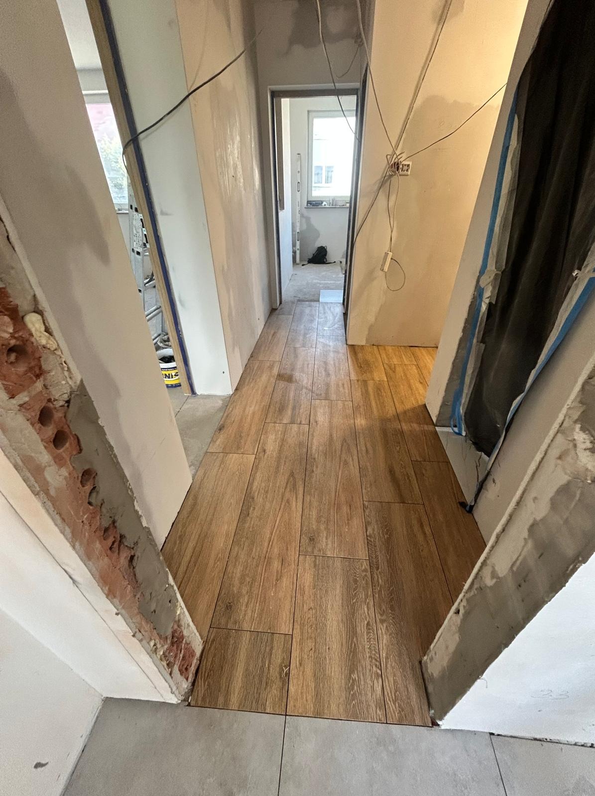 Korytarz w trakcie remontu: ułożona drewnopodobna podłoga z płytek, ściany w stanie surowym, widoczne kable i elementy konstrukcyjne. Perspektywa z wnętrza pomieszczenia.