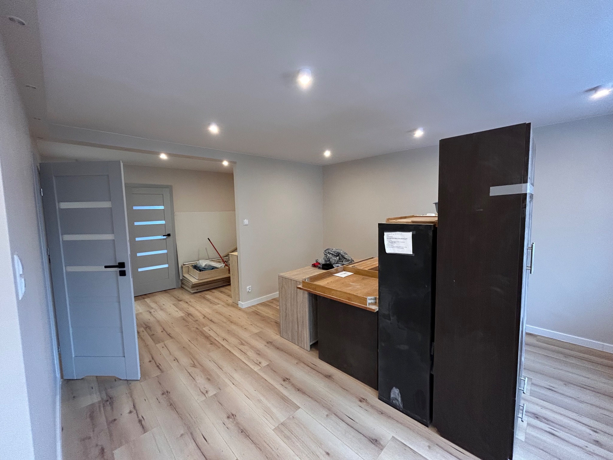 Jasne panele podłogowe w nowym wnętrzu z szarymi drzwiami i świeżo pomalowanymi ścianami. Widoczne elementy wyposażenia w trakcie montażu.