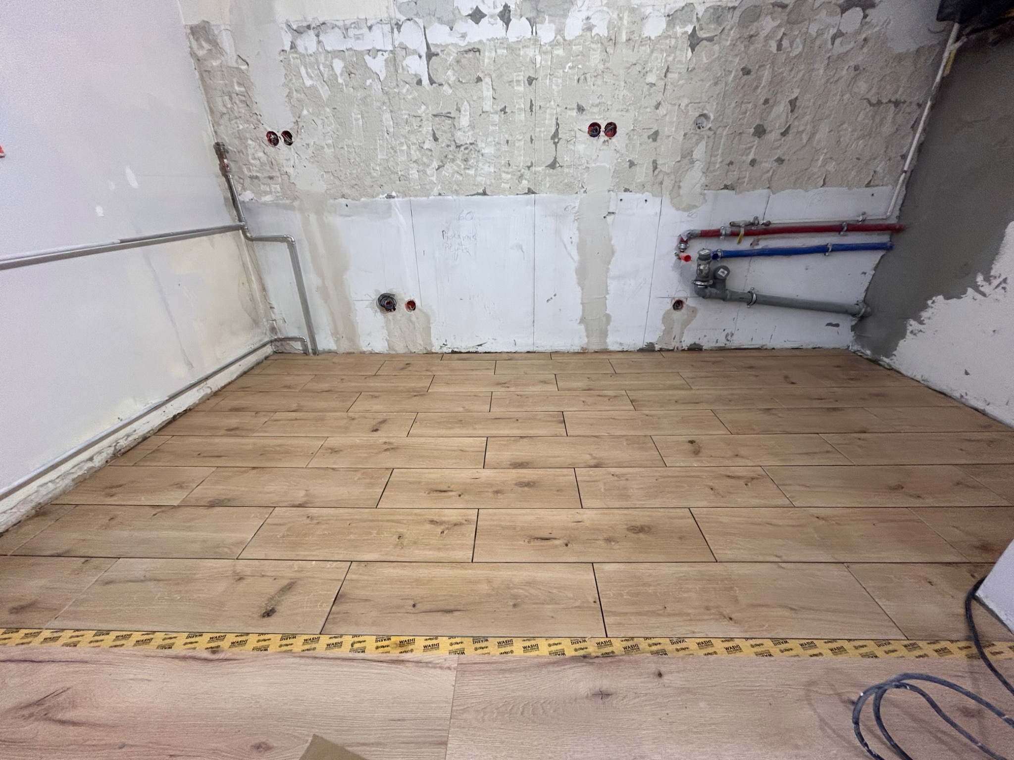 Ułożona podłoga z płytek imitujących drewno w pomieszczeniu w trakcie prac wykończeniowych. Widoczne instalacje i surowe ściany.