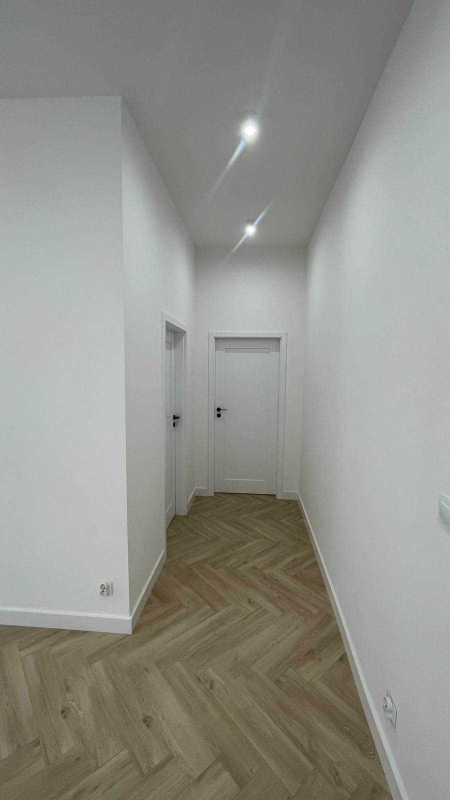 Korytarz po remoncie z parkietem ułożonym w jodełkę, białe ściany i dwoje białych drzwi. Oświetlenie punktowe w suficie. Widok z dołu.
