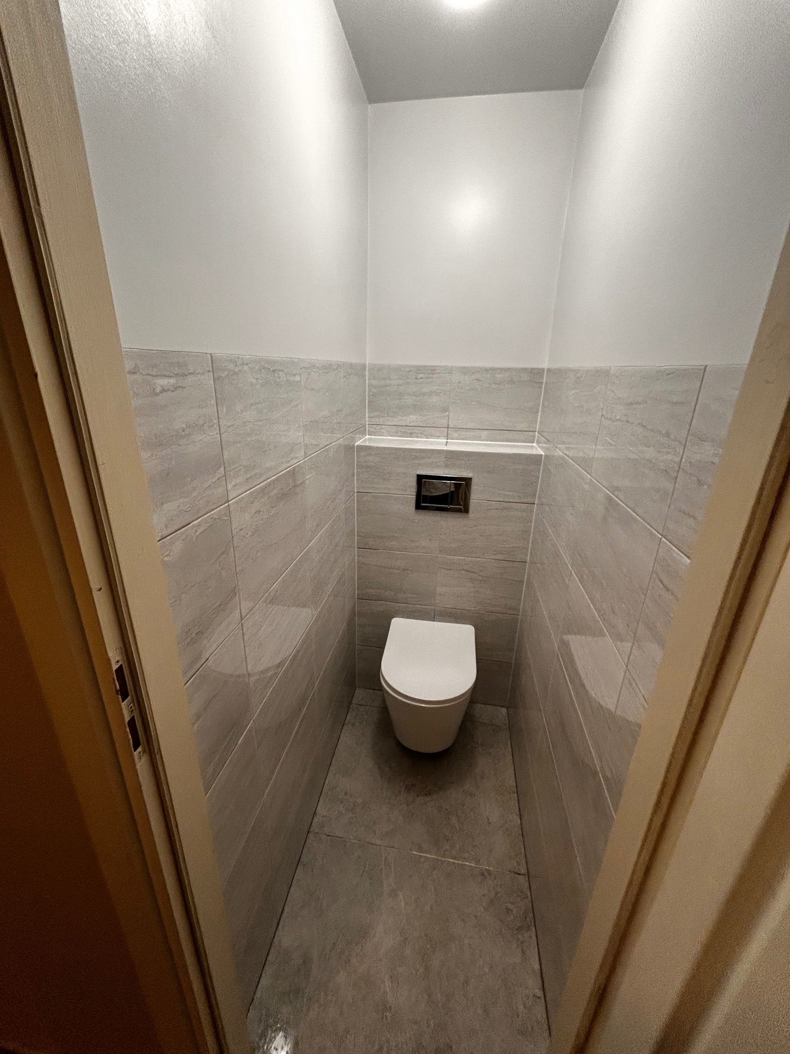 Wykończona toaleta w szarościach: płytki imitujące kamień na ścianach i podłodze, podwieszana miska WC, chromowany przycisk spłukujący. Widok zza uchylonych drzwi.