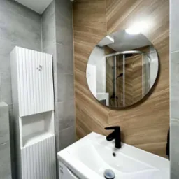 Nowoczesna łazienka z umywalką, szafką i okrągłym lustrem, w którym odbija się kabina prysznicowa. Ściany wyłożone płytkami w odcieniach szarości i drewna.