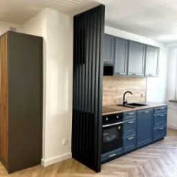 Nowoczesna kuchnia z szafkami w odcieniach szarości i niebieskiego, drewnianym blatem oraz podłogą w jodełkę. Czarny ażurowy panel oddziela przestrzeń.