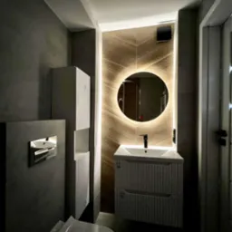 Nowoczesna łazienka z podświetlanym lustrem, umywalką z szafką w kolorze złamanej bieli i szarymi płytkami. Uwagę przykuwa drewniana ściana za lustrem.