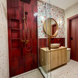 Krzysztof Stachurski Homecraft - Wykończona łazienka z kabiną prysznicową, czerwonymi płytkami i armaturą w kolorze złota. Umywalka nablatowa na drewnianej szafce, okrągłe lustro i tapeta w muszelki.