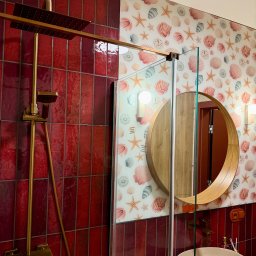 Krzysztof Stachurski Homecraft - Nowoczesna łazienka z czerwonymi płytkami, złotą armaturą i okrągłym lustrem na ścianie w muszelki. Kabina prysznicowa ze szklanymi drzwiami.