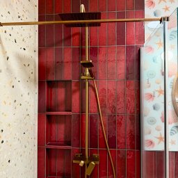 Krzysztof Stachurski Homecraft - Nowoczesna kabina prysznicowa z czerwoną, błyszczącą glazurą ułożoną pionowo, złotą armaturą i półkami wnękowymi. Ściana obok wykończona płytkami lastryko.