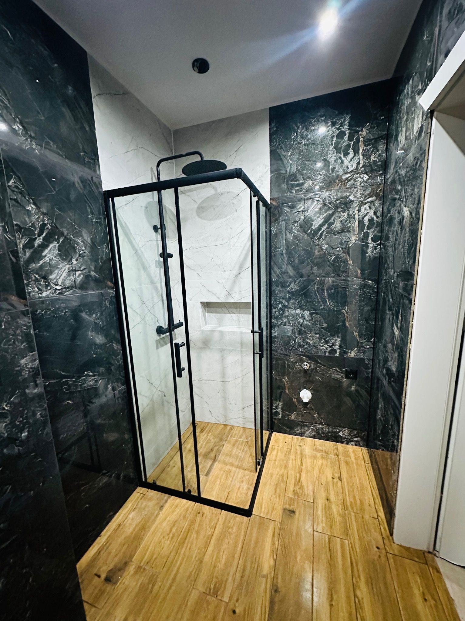 Wykończona łazienka z kabiną prysznicową narożną z czarnymi profilami, marmurowe płytki w odcieniach czerni i bieli na ścianach, drewnopodobna podłoga, minimalistyczny design.