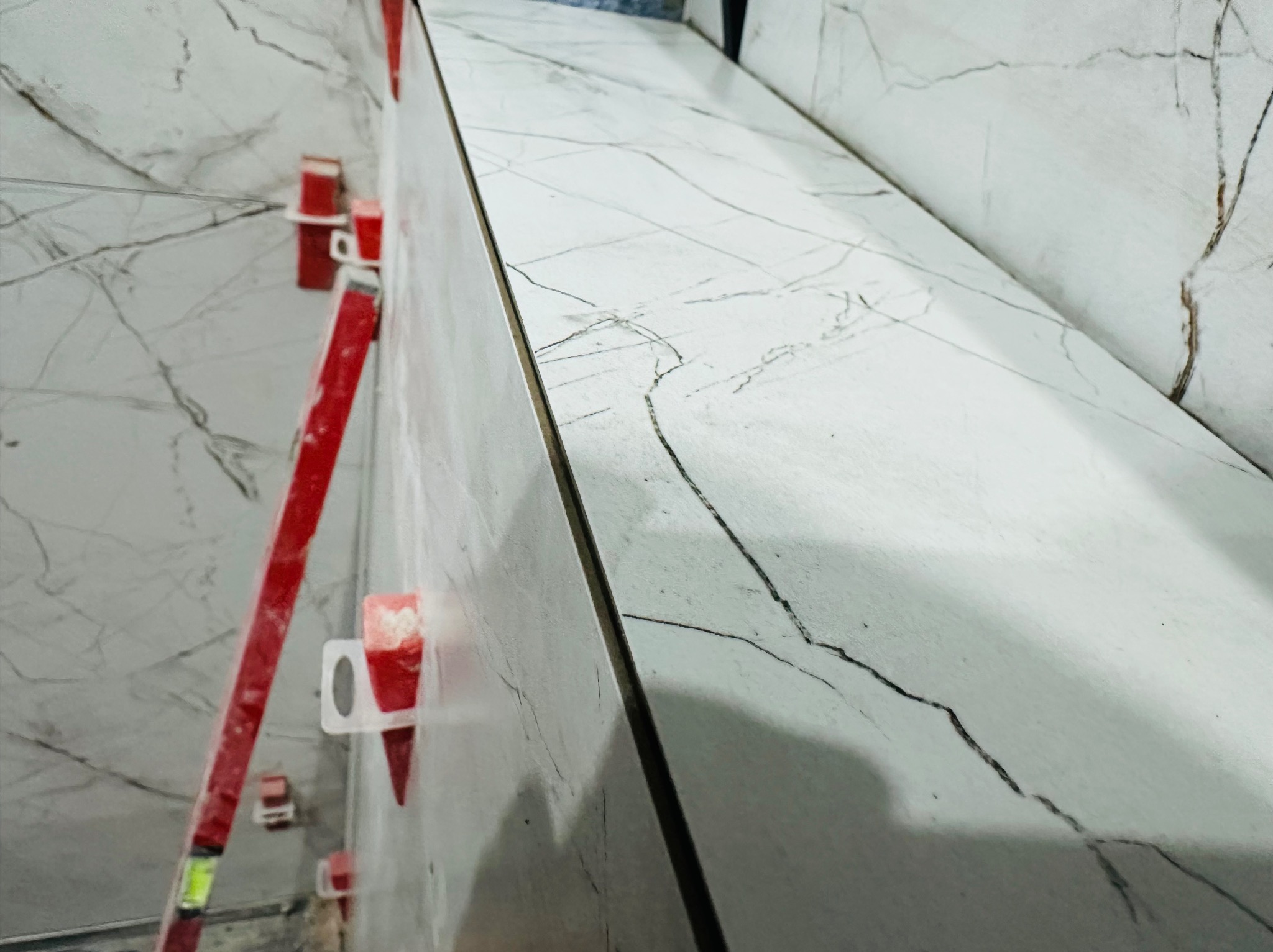 Montaż płytek imitujących marmur na ścianie z użyciem poziomicy i klipsów poziomujących, widoczne ciemne żyłki na jasnym tle.