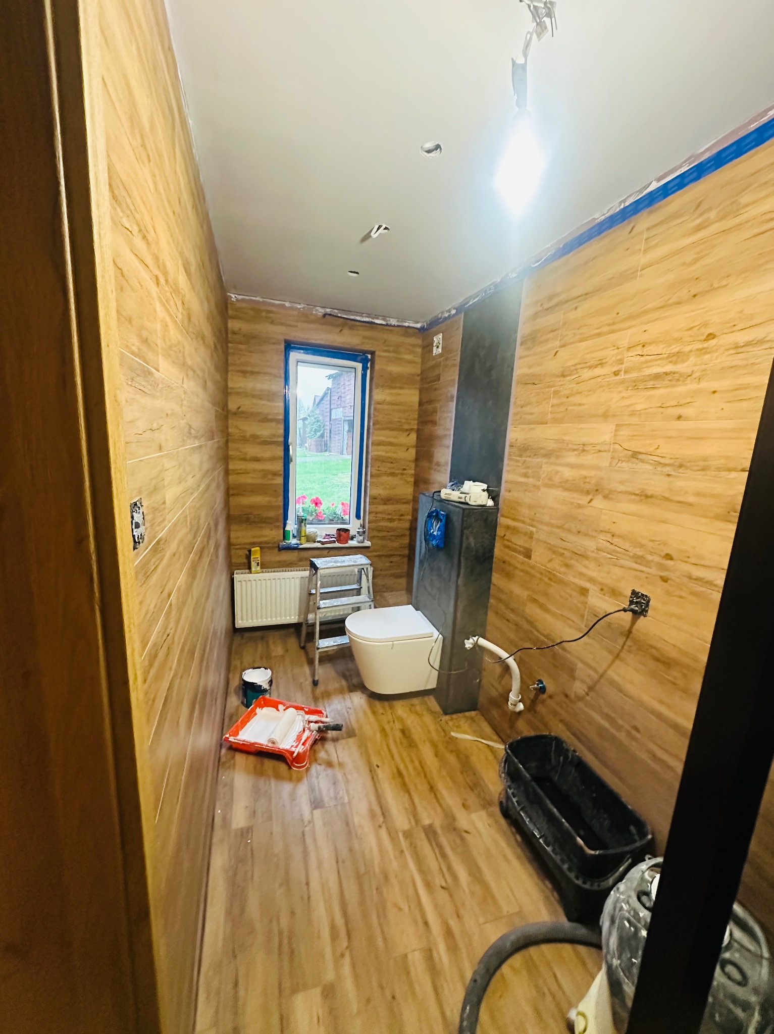 Wnętrze łazienki w trakcie prac wykończeniowych: drewnopodobne płytki na ścianach i podłodze, toaleta, drabinka, okno z widokiem na ogród, akcesoria malarskie i wiadro na podłodze, zabezpieczone...