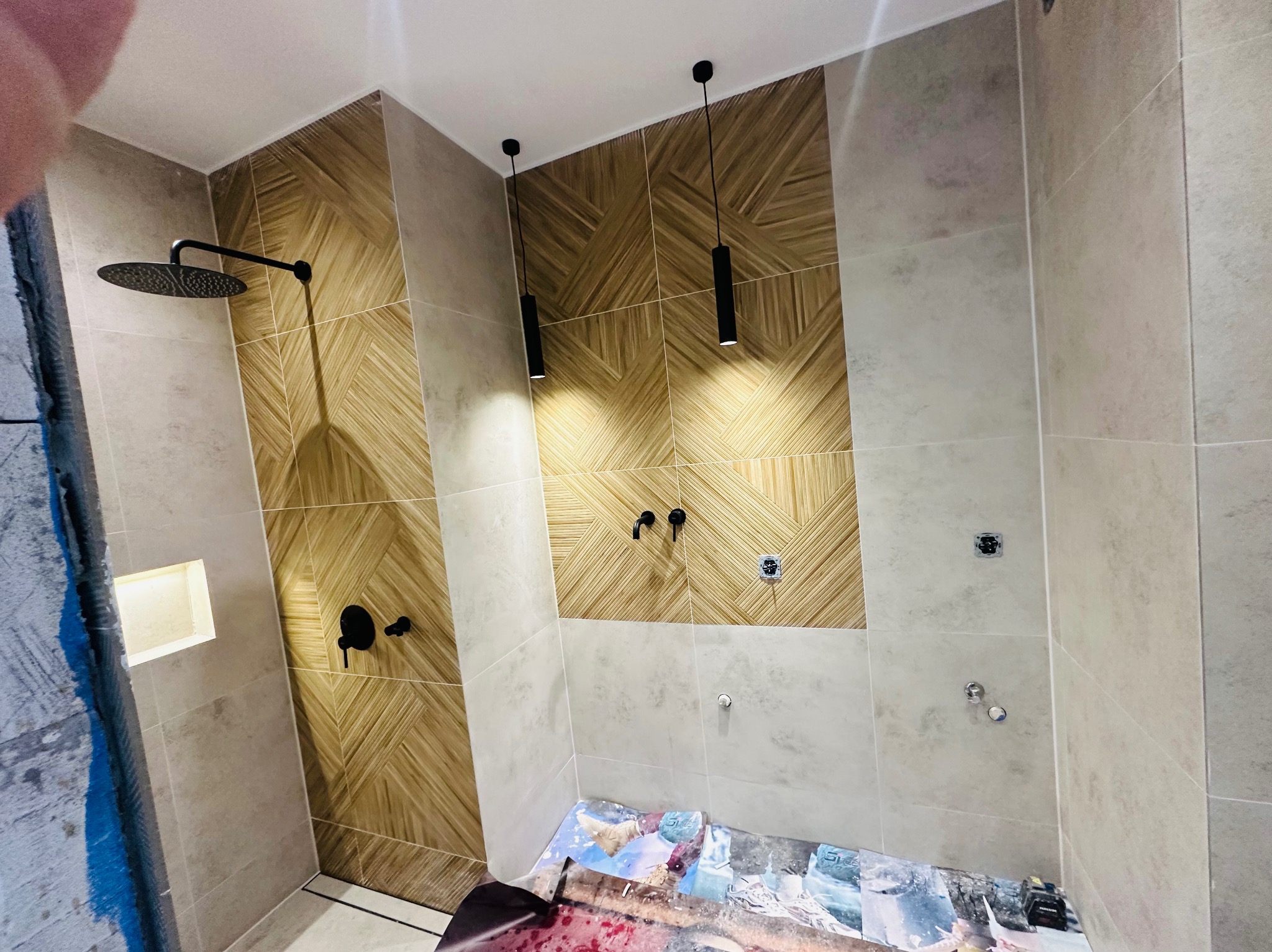 Nowoczesna łazienka w trakcie wykańczania: widoczne płytki imitujące drewno ułożone w geometryczny wzór, czarna deszczownica, minimalistyczne lampy wiszące i zabezpieczona podłoga.