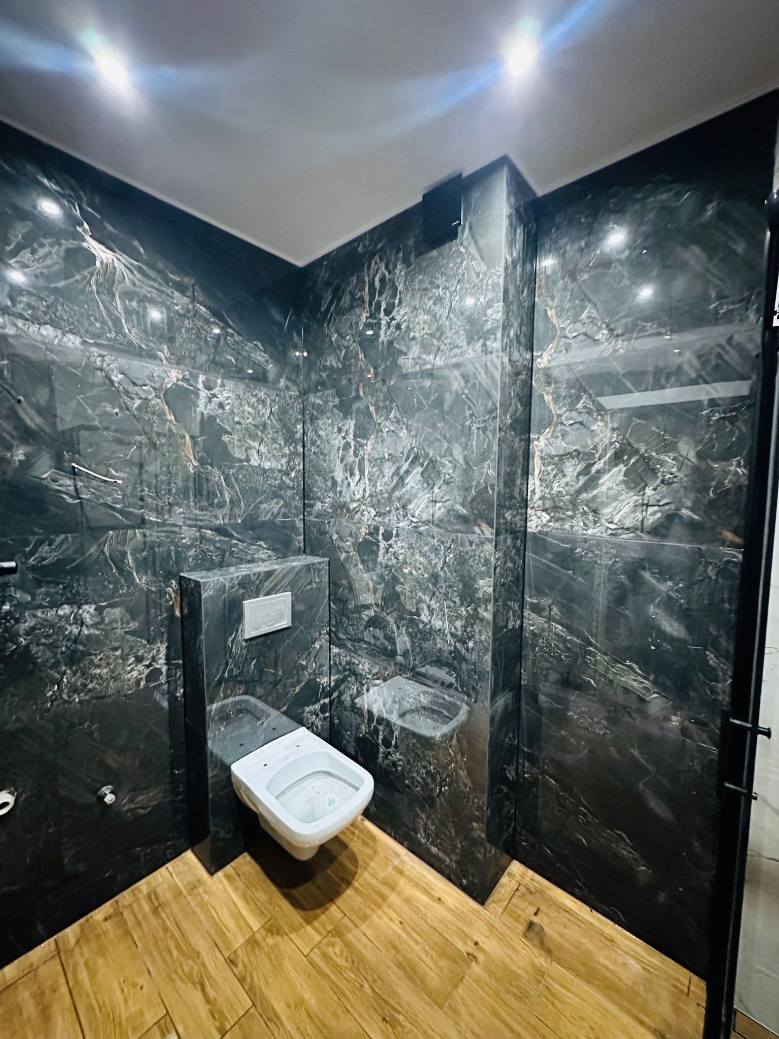 Nowoczesna łazienka z czarnymi płytkami imitującymi marmur na ścianach i podwieszaną toaletą oraz drewnopodobną podłogą, oświetlona punktowymi lampami sufitowymi.