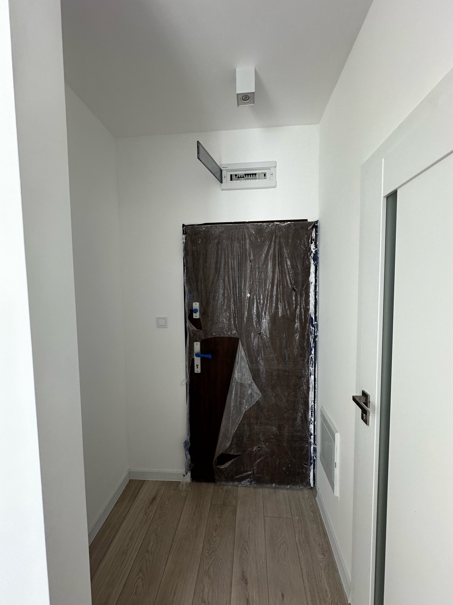 Korytarz w trakcie zabezpieczania przed pracami wykończeniowymi, drzwi wejściowe osłonięte folią malarską, widoczna skrzynka elektryczna na ścianie, podłoga pokryta panelami.