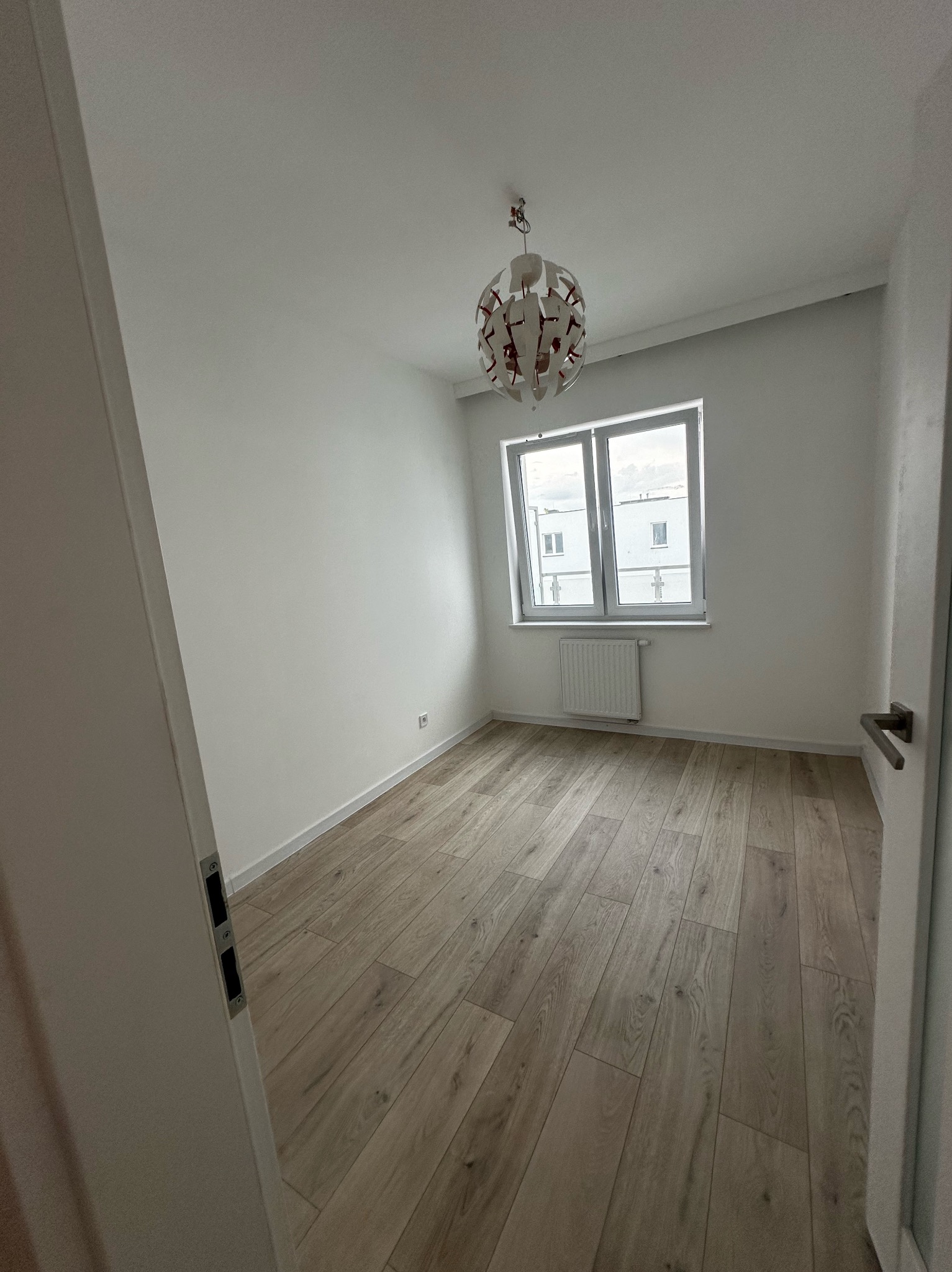 Pusty pokój z drewnianą podłogą, białymi ścianami, grzejnikiem pod oknem i nowoczesną lampą wiszącą, widok zza uchylonych drzwi.
