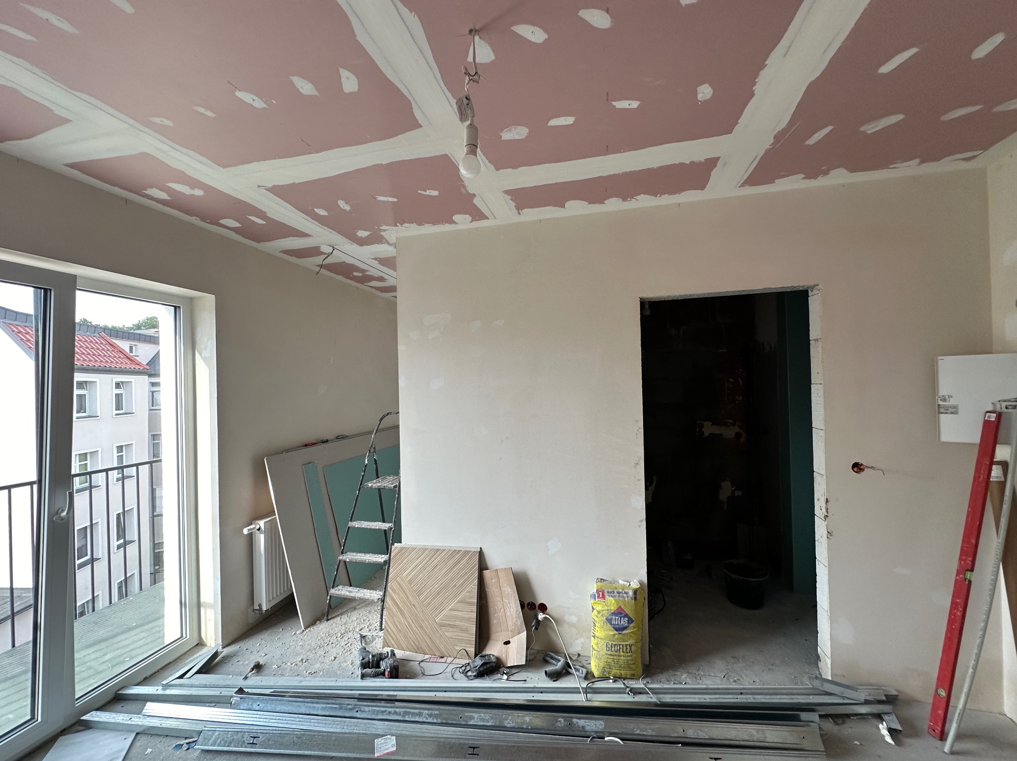 Wnętrze w trakcie remontu: sufit z płyt kartonowo-gipsowych z widocznymi łączeniami, nieotynkowane ściany, metalowe profile na podłodze, drabina, płyty gipsowe i worki z zaprawą budowlaną.