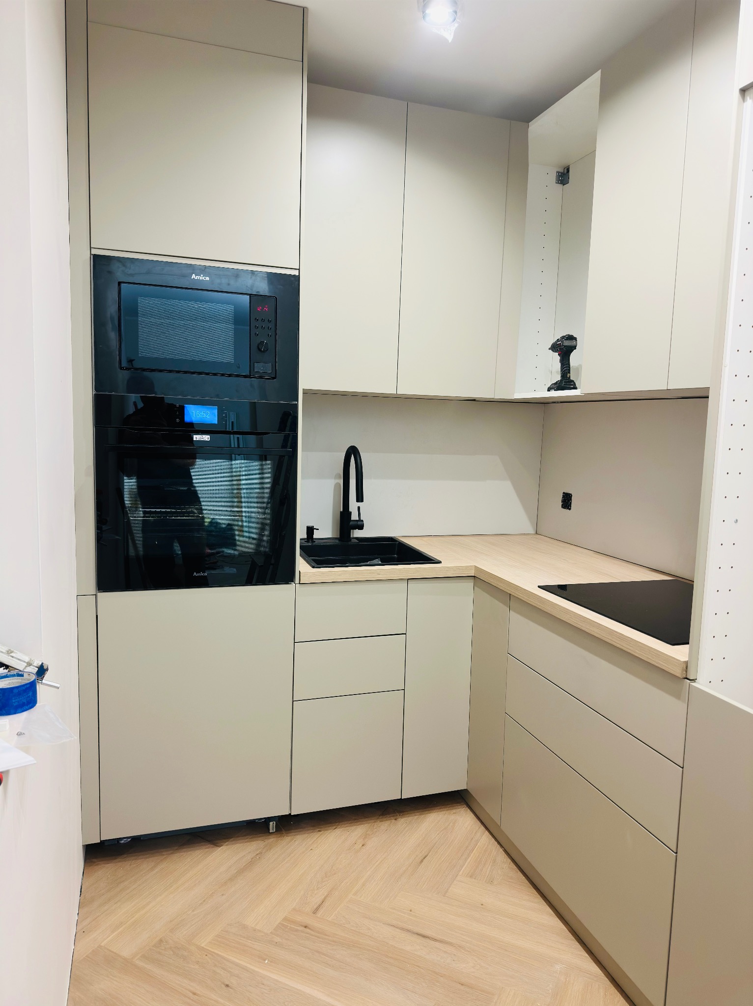 Nowoczesna kuchnia w Słupsku z beżowymi szafkami, czarnym zlewem i płytą indukcyjną, oraz parkietem ułożonym w jodełkę, w trakcie montażu widoczna wiertarka na blacie.