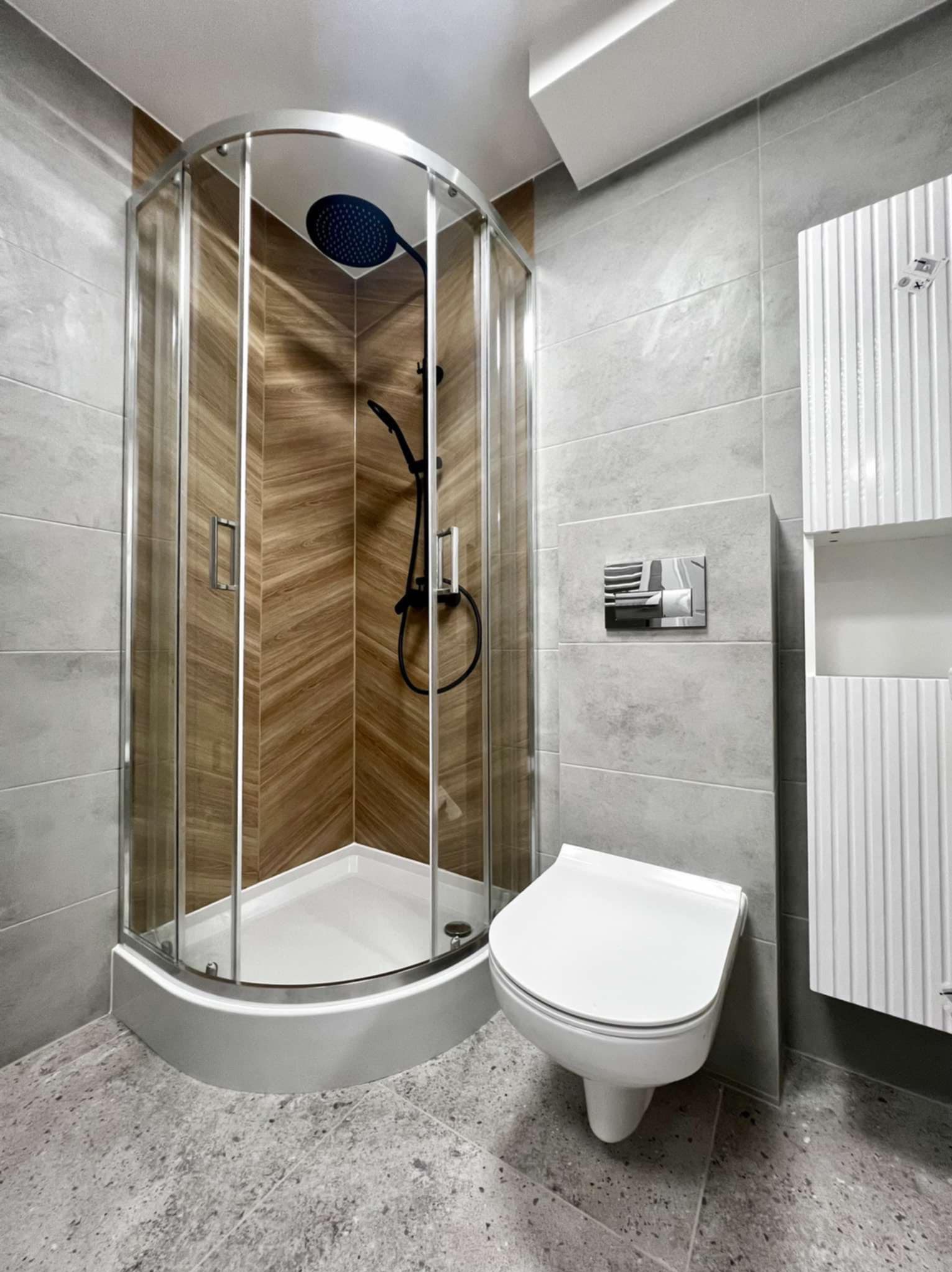 Nowoczesna łazienka z kabiną prysznicową narożną, wykończona szarą płytką i drewnopodobną okładziną w kabinie. Uwagę zwraca czarna armatura i podwieszana toaleta.