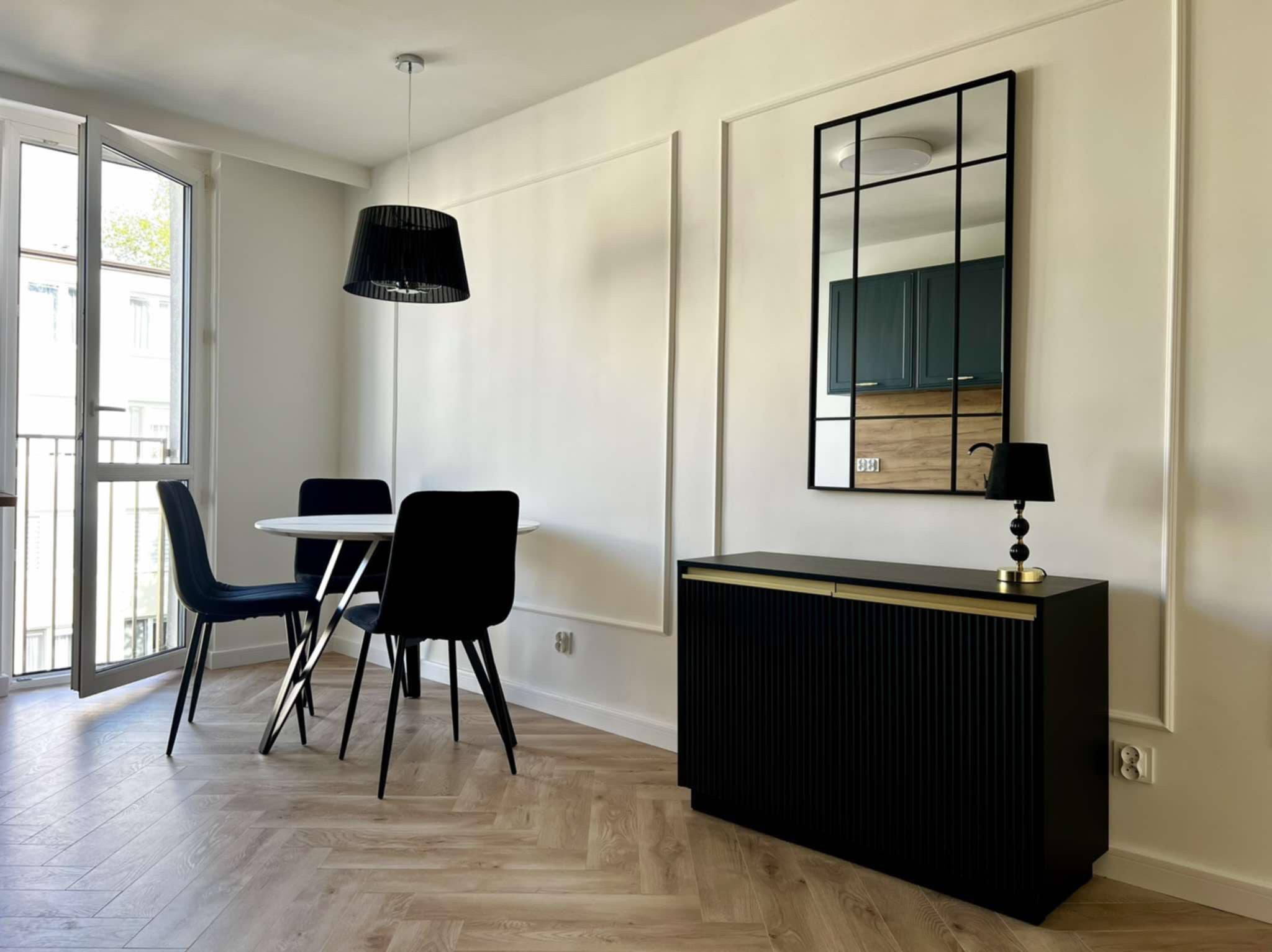 Jasne wnętrze z parkietem w jodełkę: okrągły stół z krzesłami, czarna komoda z lustrem w stylu industrialnym. Minimalistyczny design z dekoracyjnymi listwami.