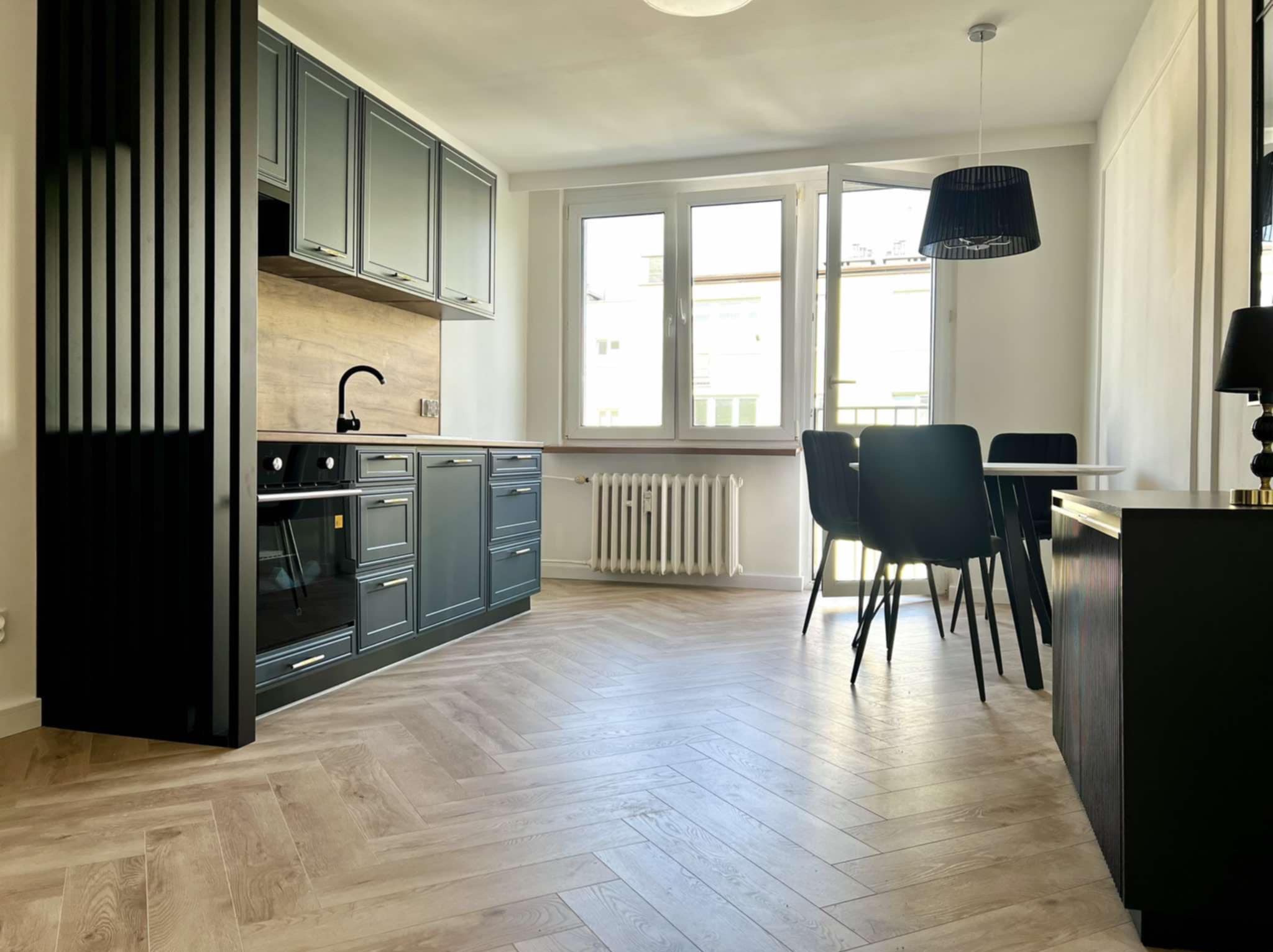 Nowoczesna kuchnia z szarymi szafkami, drewnianym blatem i parkietem w jodełkę. Widoczne czarne krzesła i lampa. Duże okno wpuszcza naturalne światło.