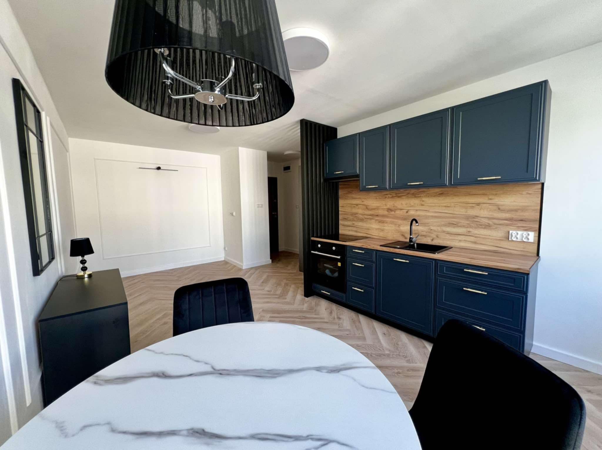 Niebieska zabudowa kuchenna z drewnianym blatem i panelami, widoczna z perspektywy stołu z marmurowym blatem i czarnymi krzesłami w nowoczesnym wnętrzu.