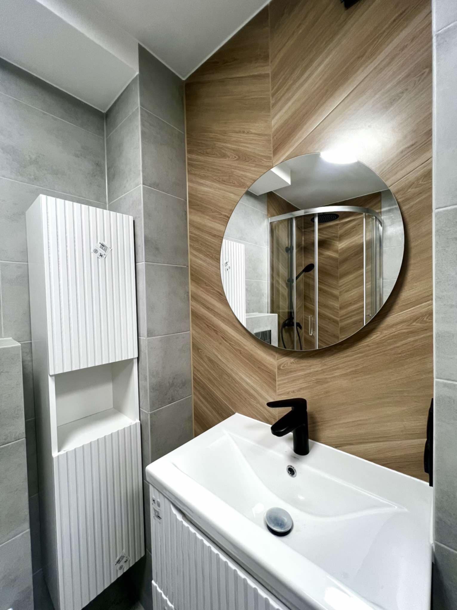 Nowoczesna łazienka z umywalką, szafką i okrągłym lustrem, w którym odbija się kabina prysznicowa. Ściany wyłożone płytkami w odcieniach szarości i drewna.