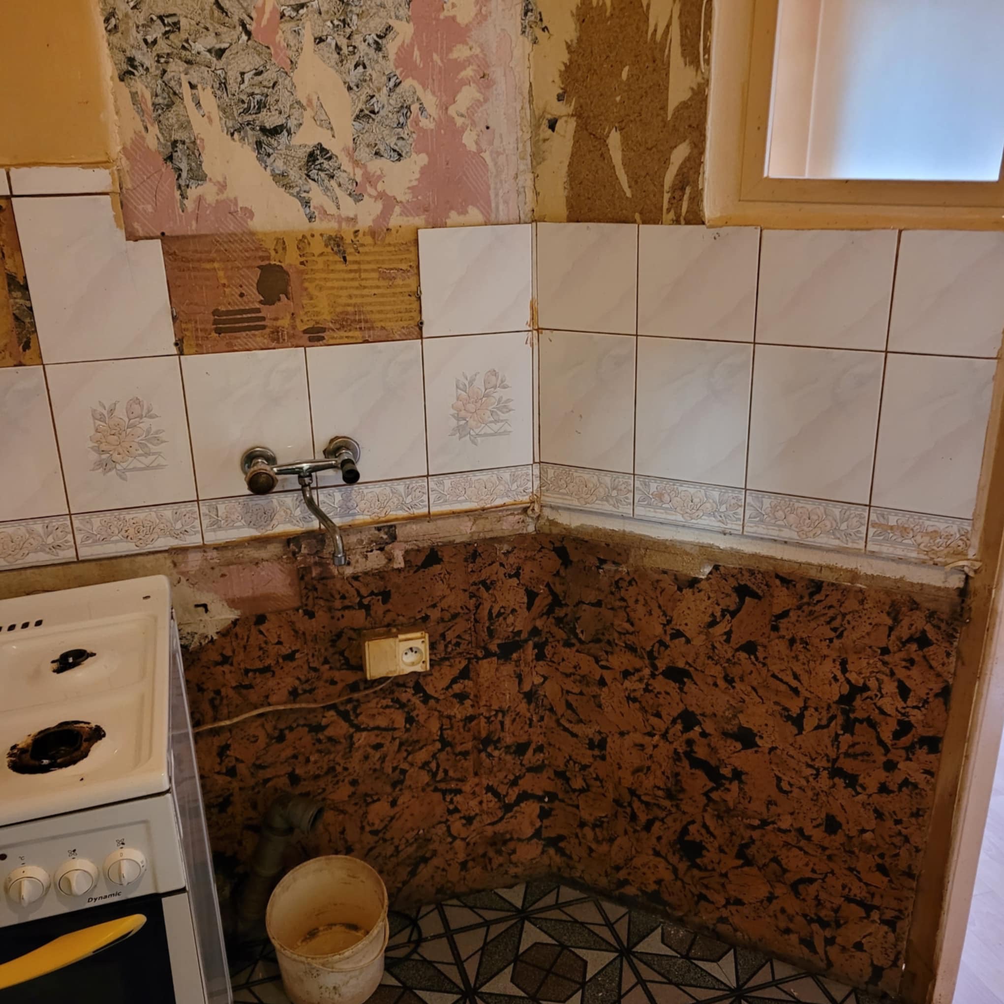 Fragment kuchni w trakcie demontażu płytek z motywem kwiatowym. Widoczne ślady po kleju, stara instalacja elektryczna i fragment kuchenki.