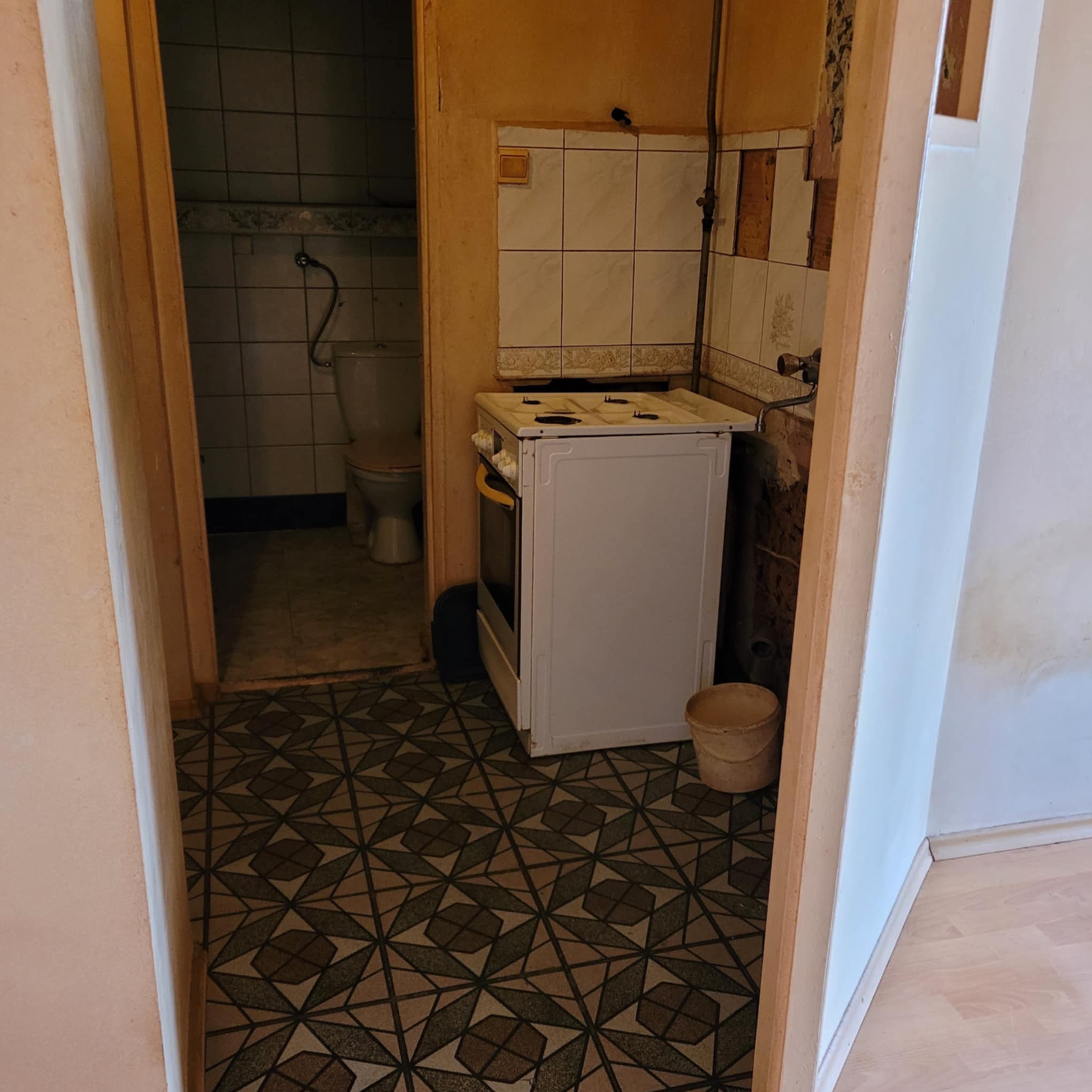 Wnętrze przed remontem: widok na starą kuchnię z białą kuchenką i fragment łazienki z toaletą, przez futrynę drzwi z wzorzystą podłogą.