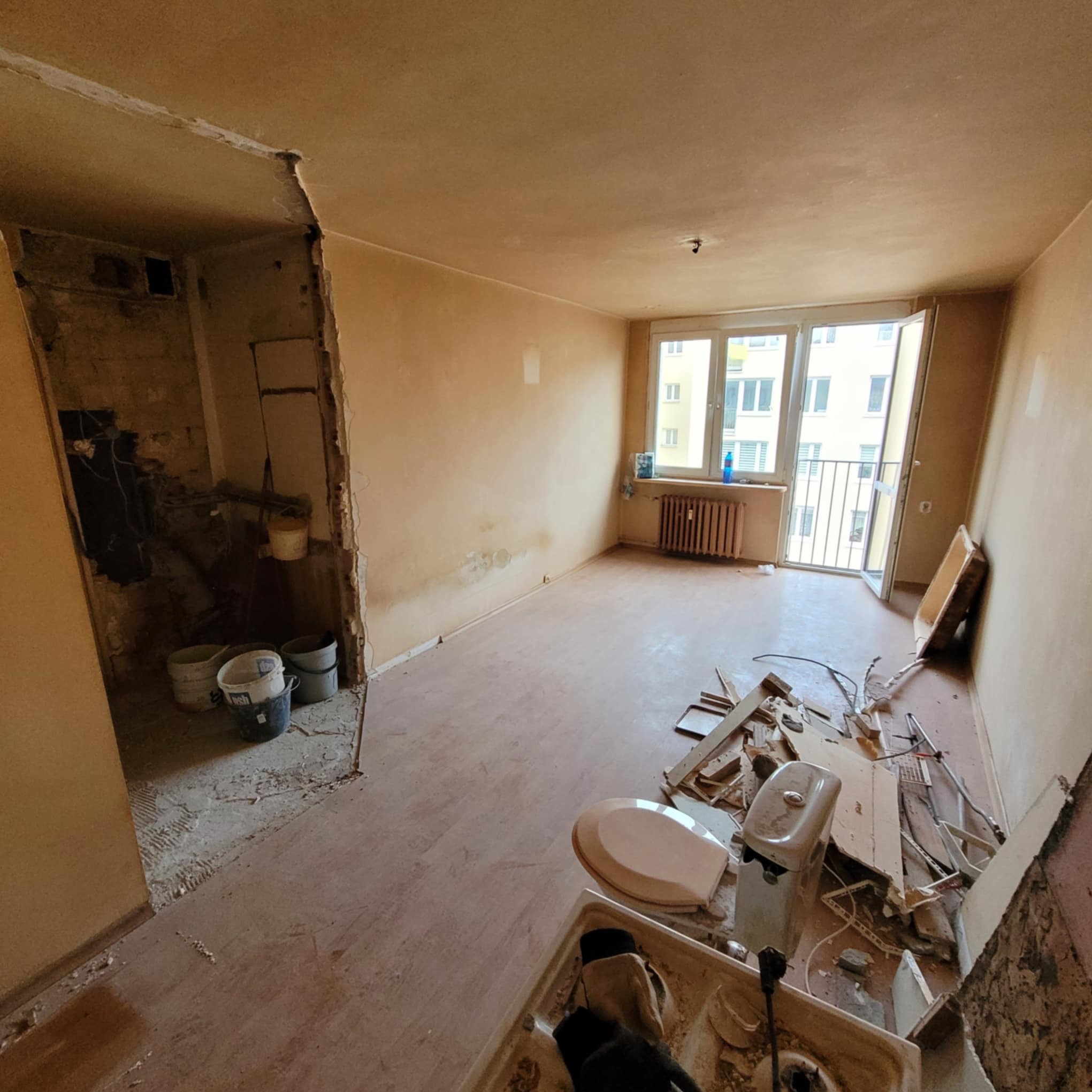 Wnętrze mieszkania w trakcie demontażu: widoczne resztki wyposażenia łazienki, odsłonięte ściany i podłogi, okno balkonowe z widokiem na budynek.