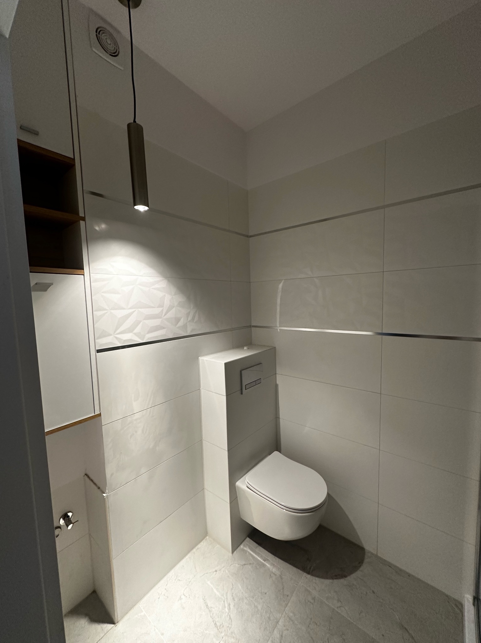 Nowoczesna toaleta w jasnej tonacji. Geometryczne płytki, podwieszana miska WC, minimalistyczna lampa. Estetyczne wykończenie wnętrza.