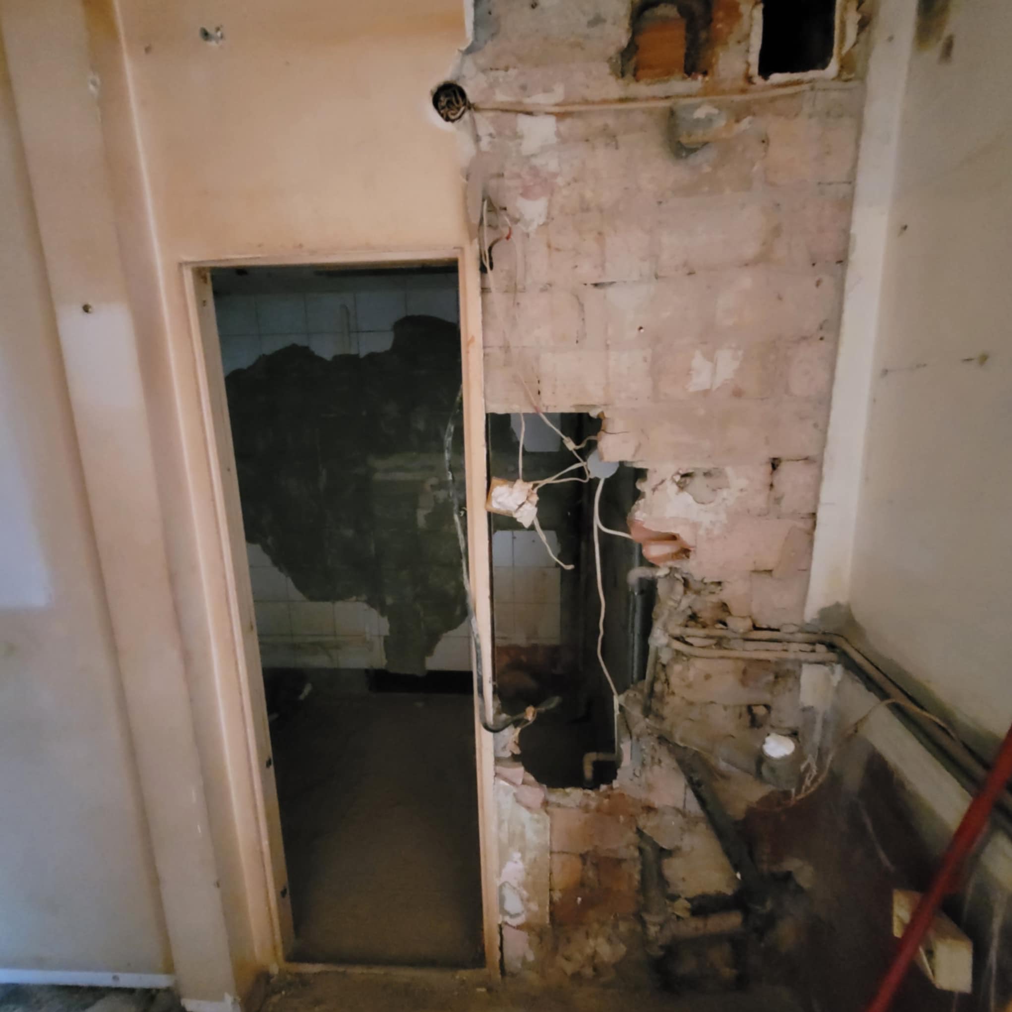 Demontaż ściany: odsłonięta cegła i instalacje w trakcie prac przygotowawczych do remontu. Widoczne przewody elektryczne i fragmenty starej łazienki w tle.