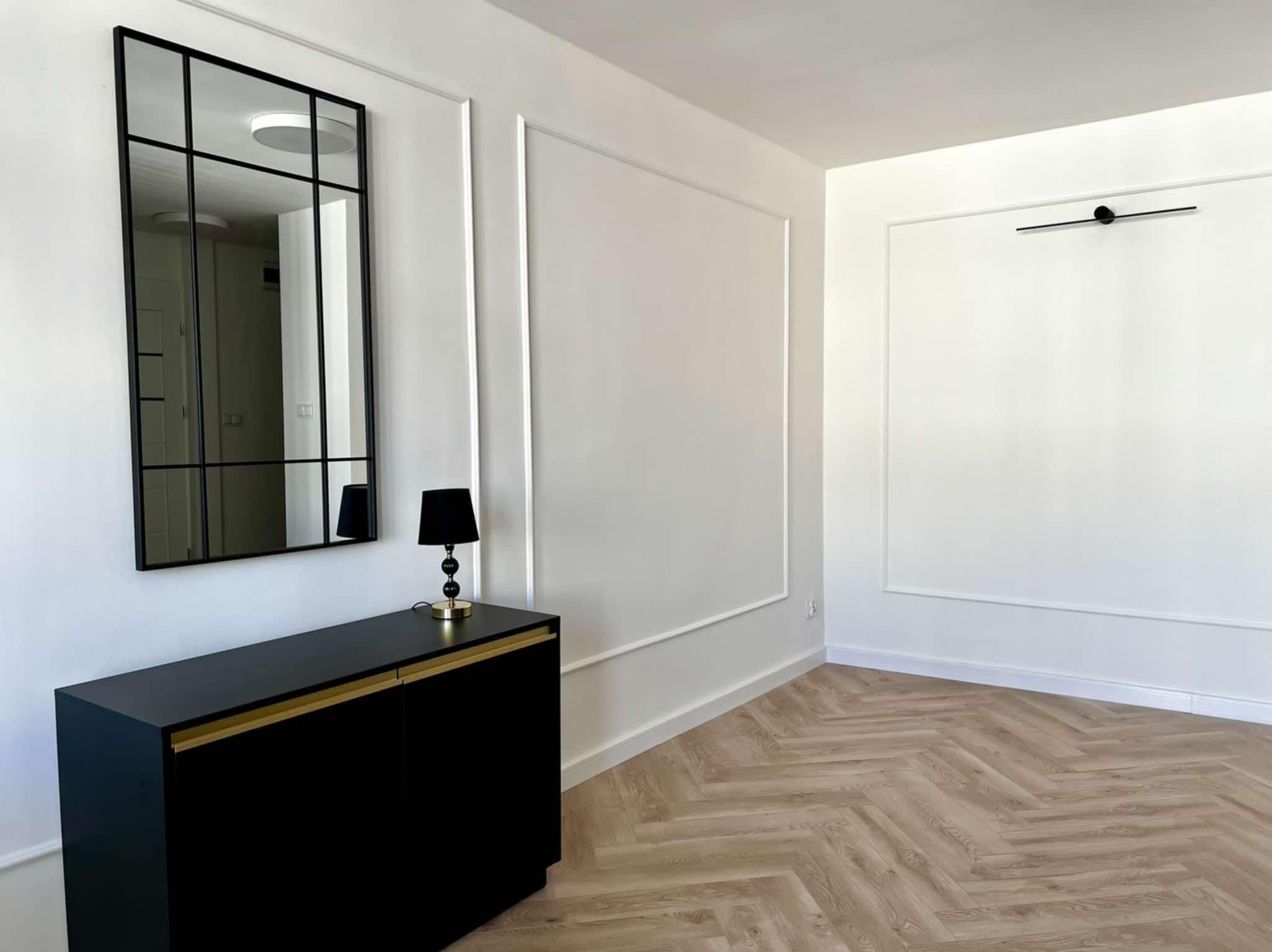 Minimalistyczne wnętrze: czarna komoda z lustrem w ramie, lampka nocna i parkiet w jodełkę. Ściany białe z dekoracyjnymi listwami.