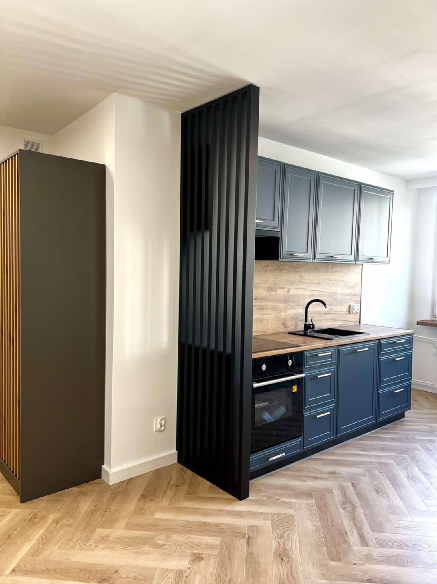 Nowoczesna kuchnia z szafkami w odcieniach szarości i niebieskiego, drewnianym blatem oraz podłogą w jodełkę. Czarny ażurowy panel oddziela przestrzeń.