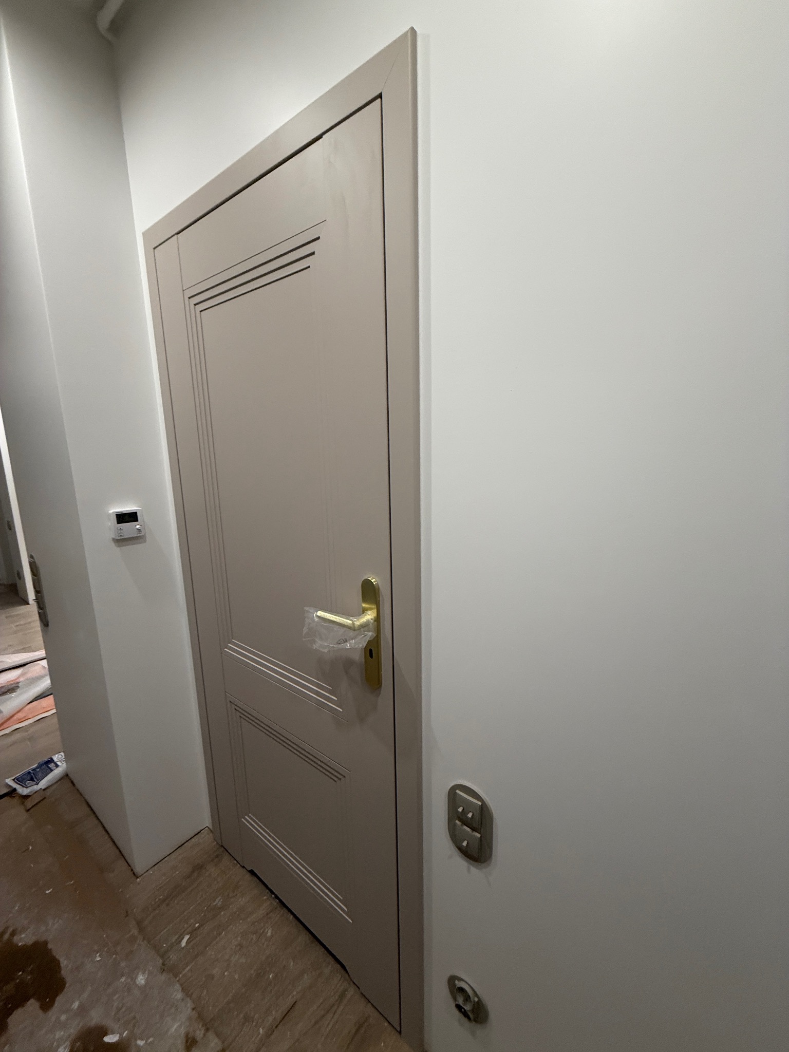 Beżowe drzwi wewnętrzne z ozdobnymi frezami i złotą klamką, zabezpieczoną folią, w nowo wykończonym wnętrzu z białymi ścianami i drewnianą podłogą.