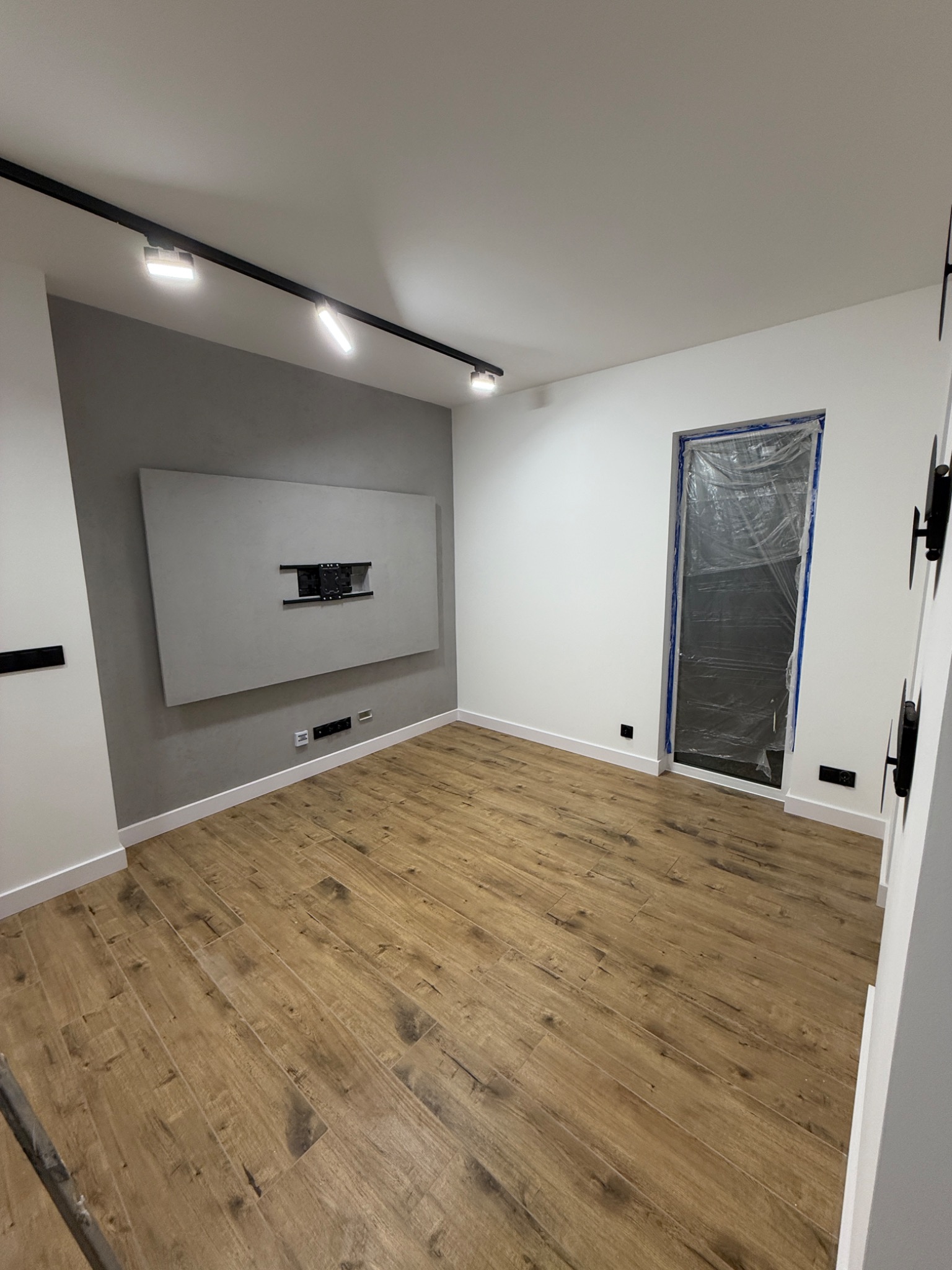 Nowoczesne wnętrze pokoju z drewnopodobną podłogą, szarą ścianą z uchwytem TV i oświetleniem szynowym. Okno zabezpieczone folią malarską.