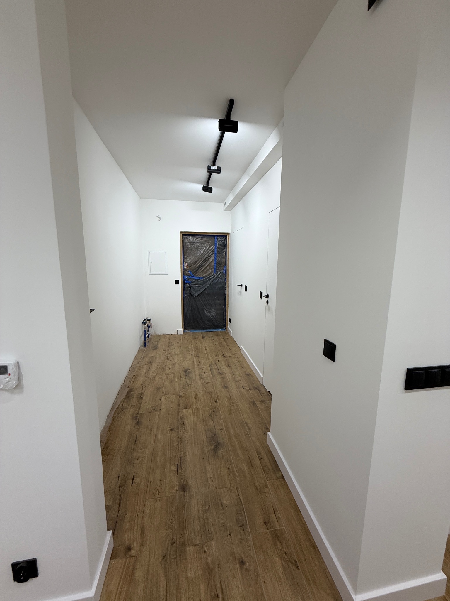 Korytarz po remoncie z drewnopodobną podłogą, białymi ścianami i czarnymi akcentami: oświetlenie, włączniki, klamki. Drzwi zabezpieczone folią malarską.