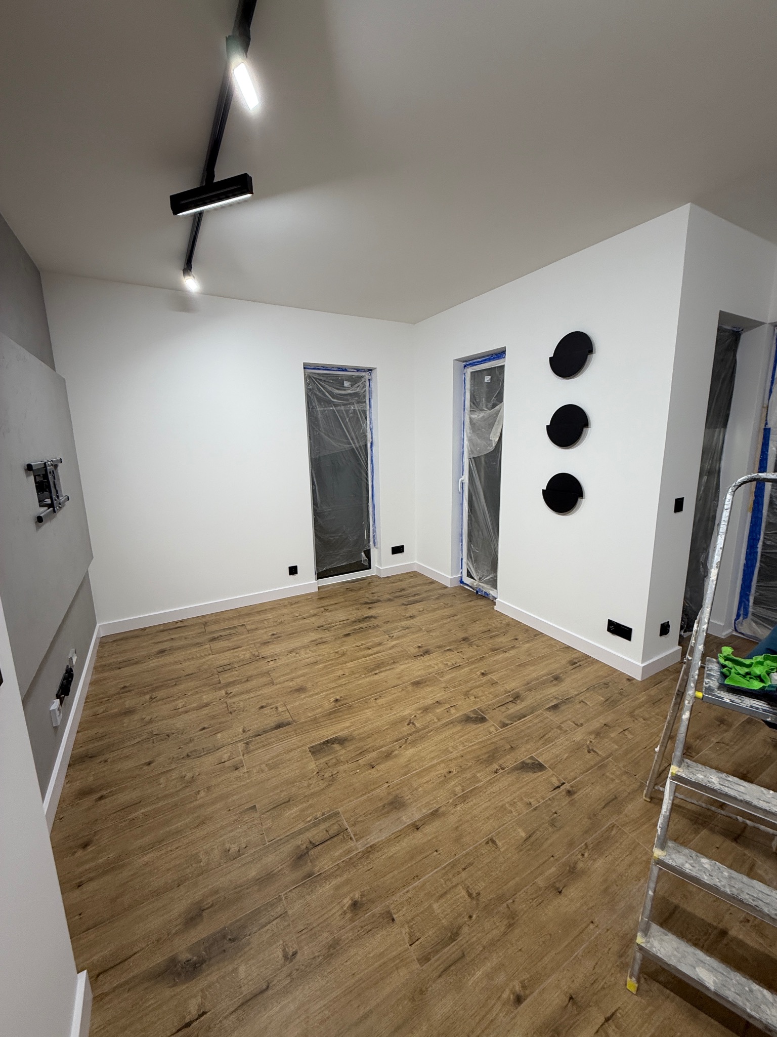 Jasne wnętrze po remoncie: drewnopodobna podłoga, białe ściany z czarnymi dekorami, nowoczesne oświetlenie szynowe. Drzwi zabezpieczone folią malarską, drabina malarska.