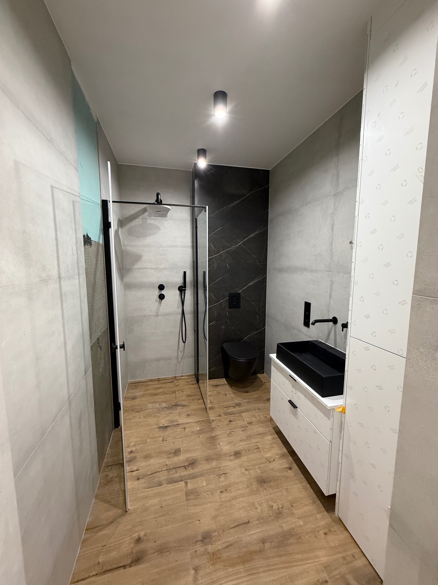 Nowoczesna łazienka z kabiną prysznicową typu walk-in, czarną armaturą i umywalką nablatową. Jasne płytki i drewnopodobna podłoga tworzą spójną całość.