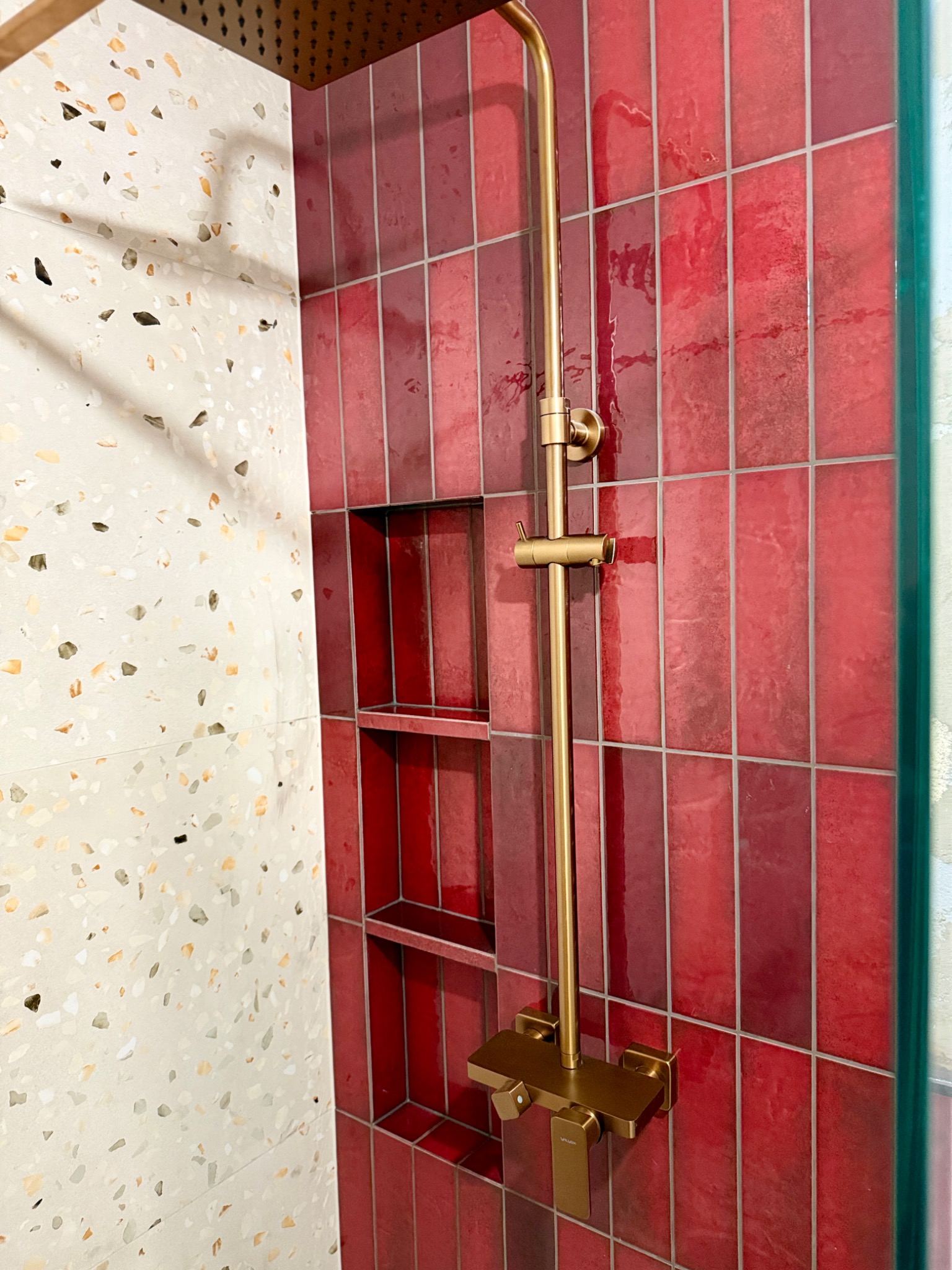 Czerwona, wąska glazura w kabinie prysznicowej z wnęką i złotą armaturą. Ściana obok wykończona płytami lastryko.