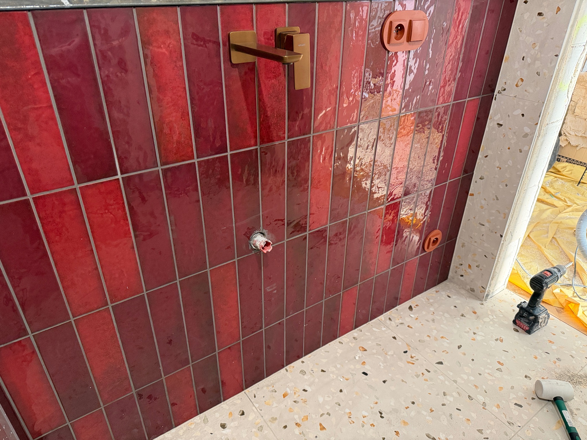 Czerwona, błyszcząca glazura ułożona pionowo na ścianie łazienki, z widoczną złotą baterią i gniazdami elektrycznymi. Na podłodze terazzo, w tle narzędzia.