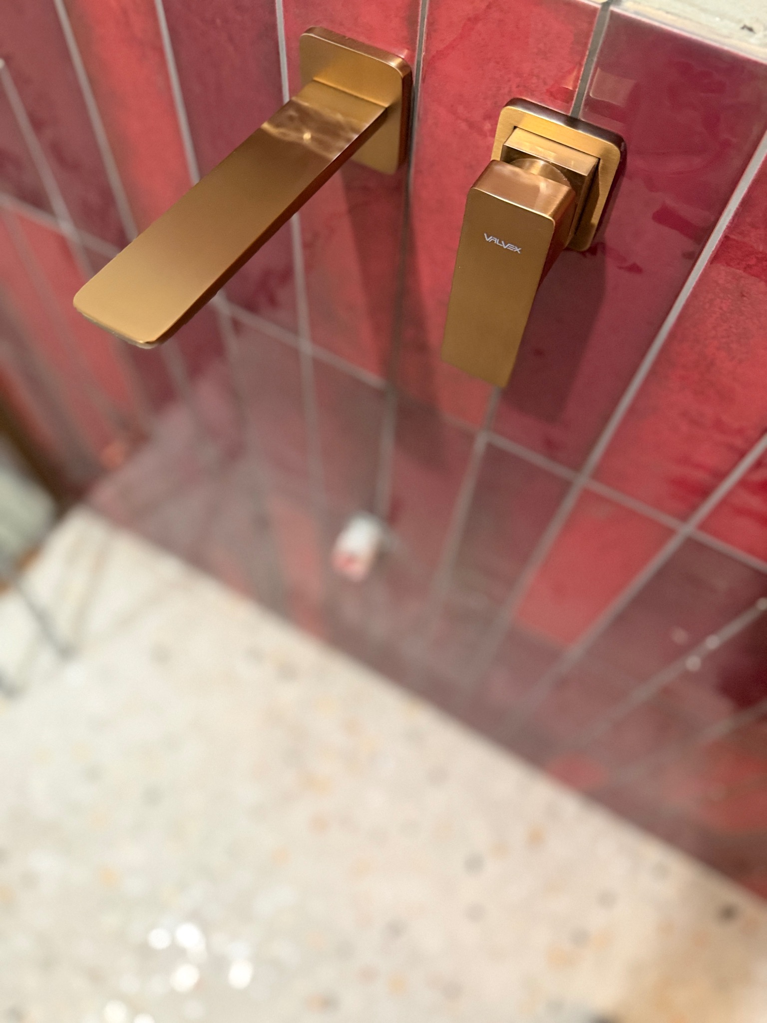 Błyszcząca, złota bateria umywalkowa marki Valvex na ścianie z czerwonych, wąskich płytek. Ujęcie z dołu, widoczny fragment umywalki. Nowoczesny design.