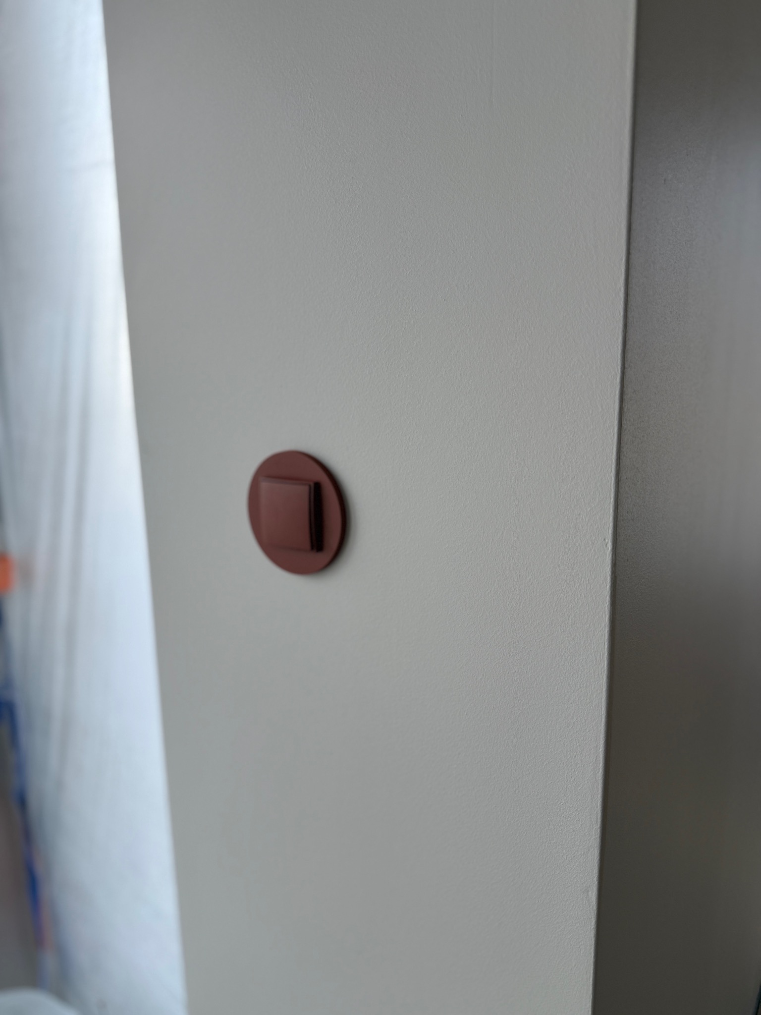 Świeżo pomalowana ściana w jasnym odcieniu z brązowym, okrągłym włącznikiem światła. Widoczne zabezpieczenia malarskie w tle.