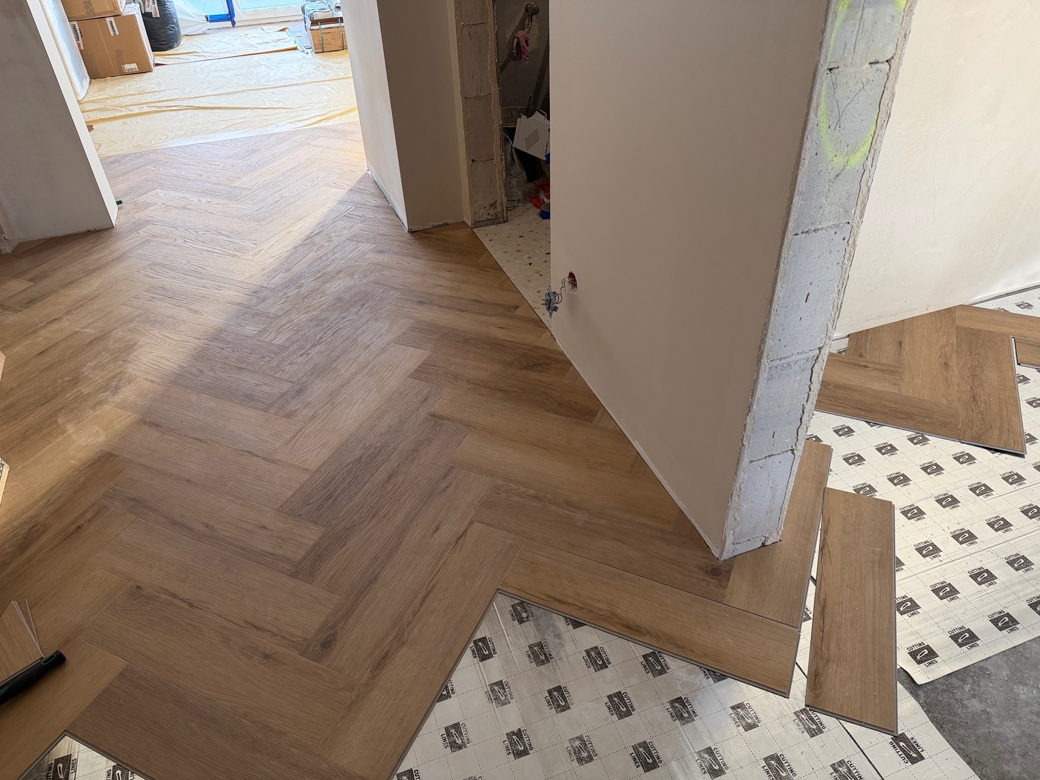 Układanie paneli podłogowych w jodełkę, widoczne podłoże z folią izolacyjną. Częściowo wykończone pomieszczenie z surowymi ścianami i instalacją elektryczną.