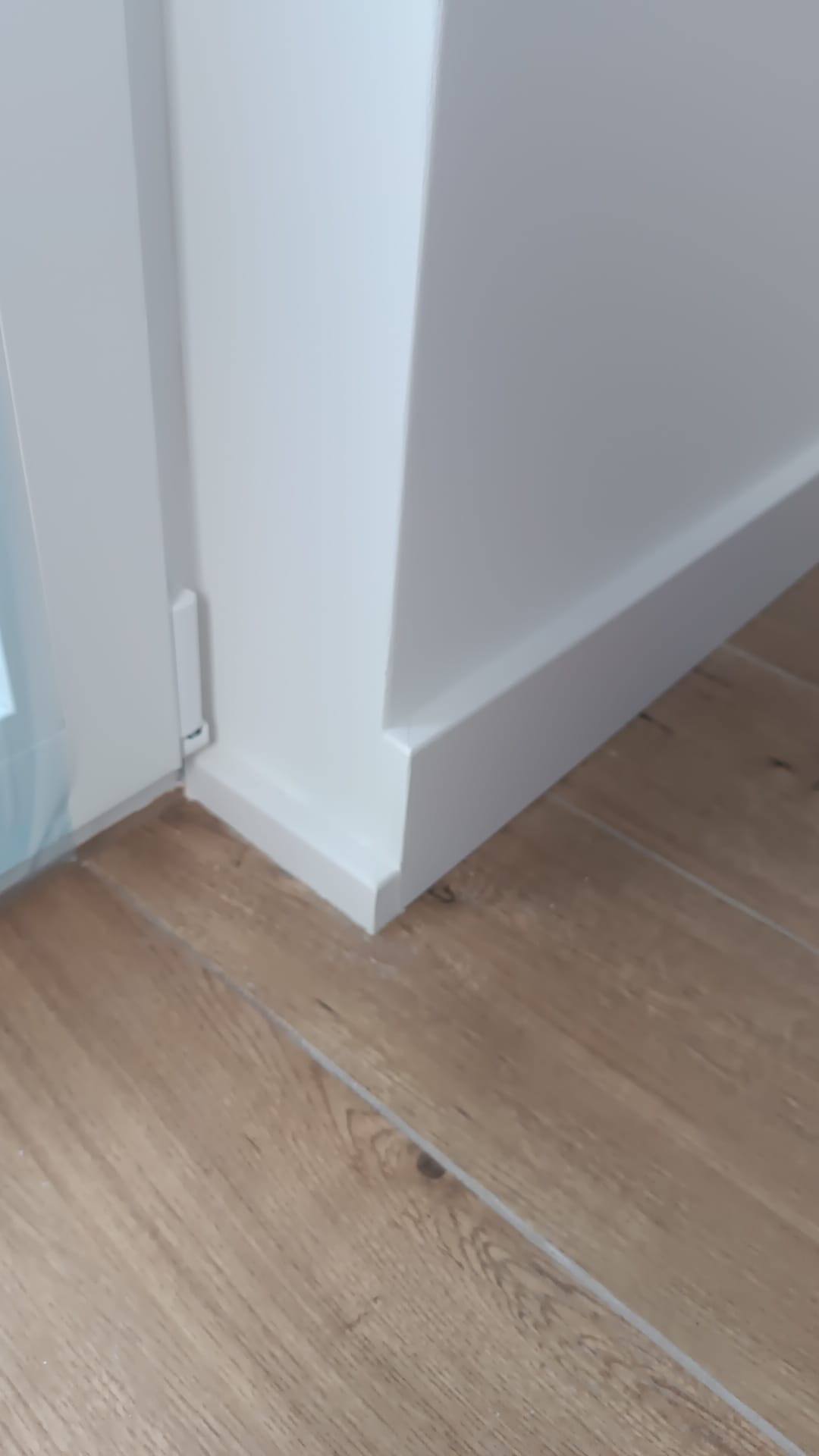 Detal wykończenia: biała listwa przypodłogowa przy ścianie i futrynie drzwiowej, na podłodze z paneli imitujących drewno. Precyzyjne łączenie elementów.