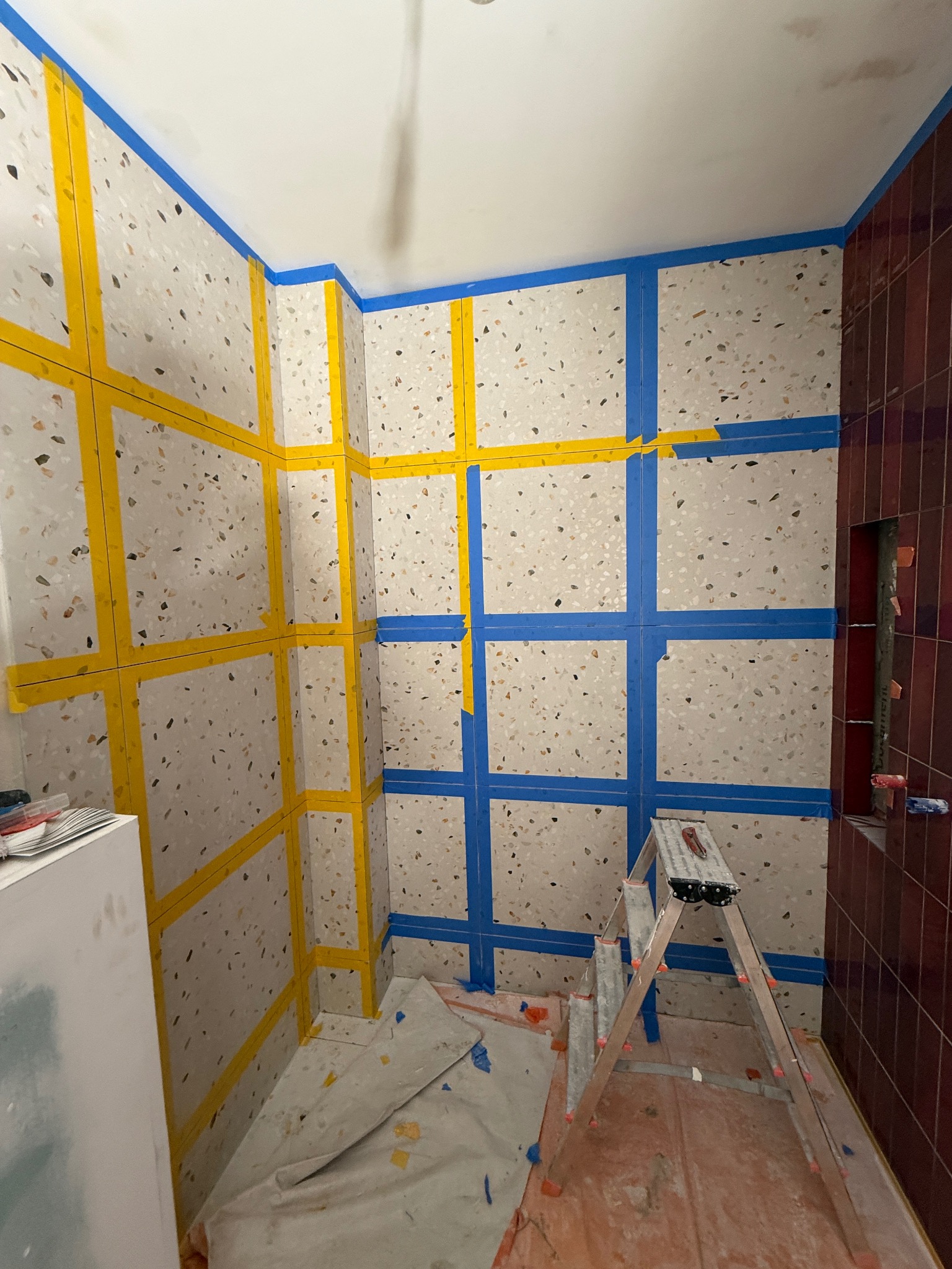 Łazienka w trakcie prac: ściany z płytek imitujących lastryko zabezpieczone taśmą malarską w kolorze żółtym i niebieskim. Widoczna drabina i zabezpieczona podłoga.