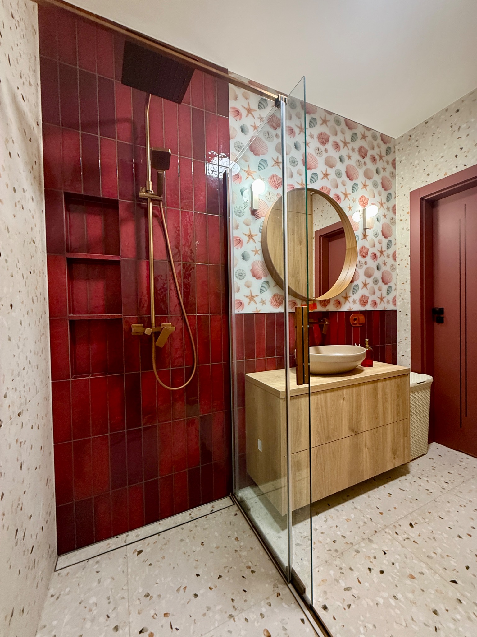 Wykończona łazienka z kabiną prysznicową, czerwonymi płytkami i armaturą w kolorze złota. Umywalka nablatowa na drewnianej szafce, okrągłe lustro i tapeta w muszelki.